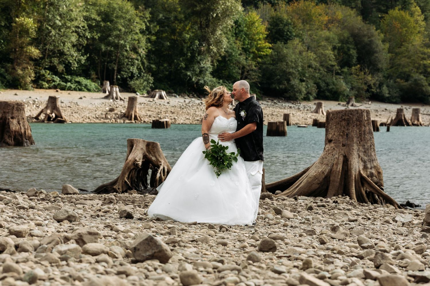 Wedding couple posing on a scenic beach with tree stumps and water in the background.