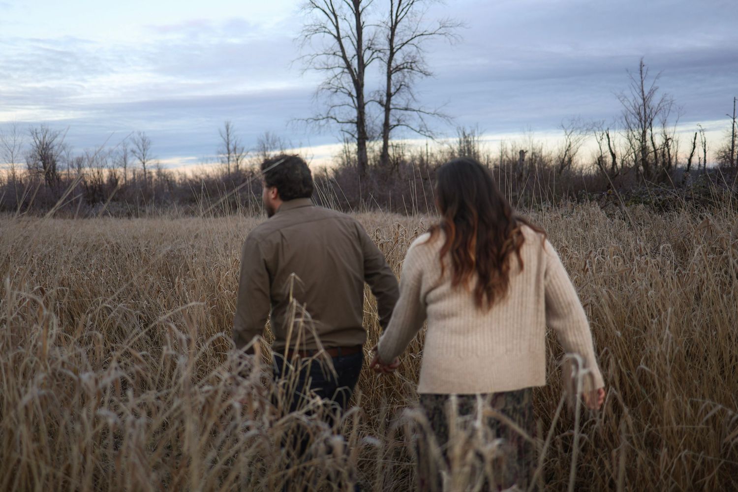 Romantic couple walking through a winter field with cloudy skies.