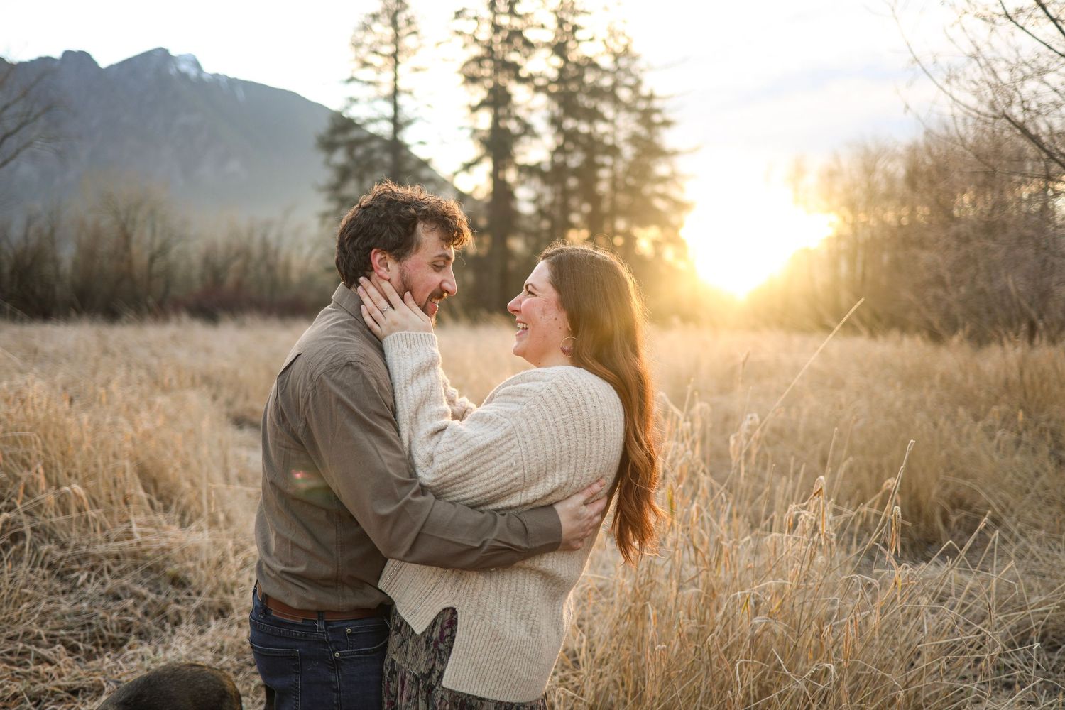 Couple sharing intimate moments in a field with evergreen trees and golden sunlight.