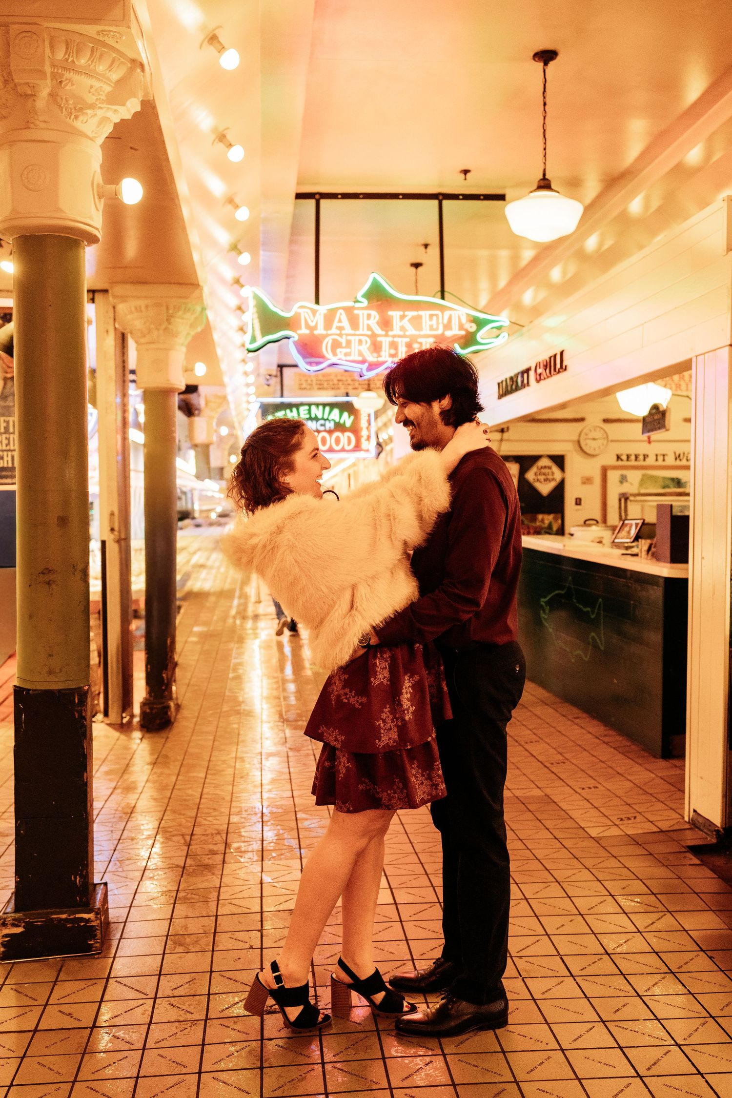 A romantic couple dancing together under warm lighting at Pike Place Market in Seattle with glowing vintage signs behind them.