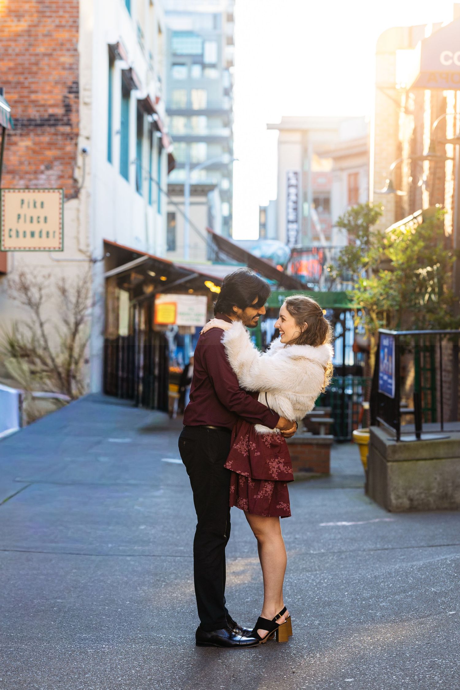 A couple embraces and kisses on a brick-lined city street with buildings in the background during golden hour.