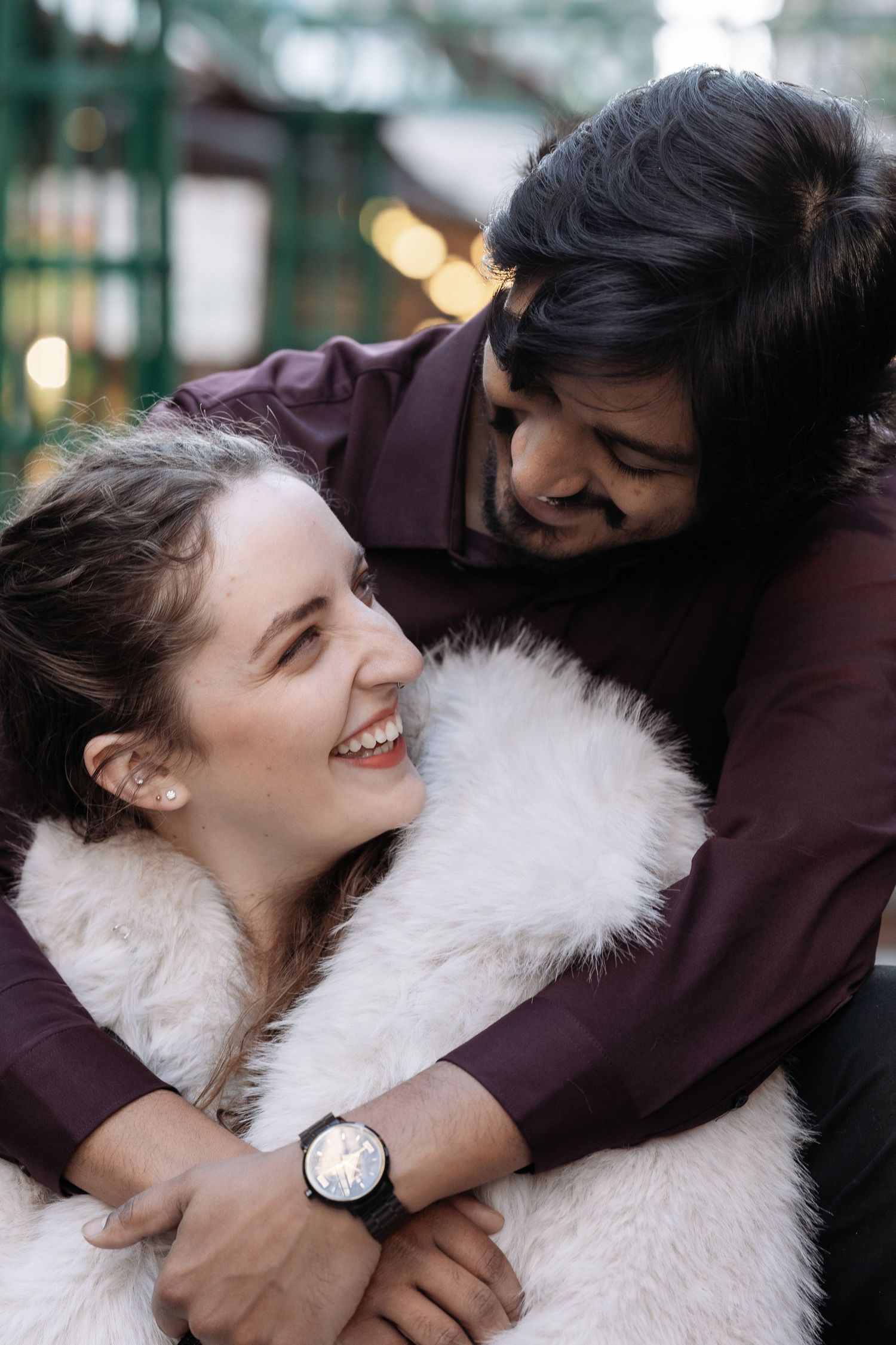 A couple shares a joyful embrace outdoors while wearing winter clothing, with soft bokeh lights in the background.
