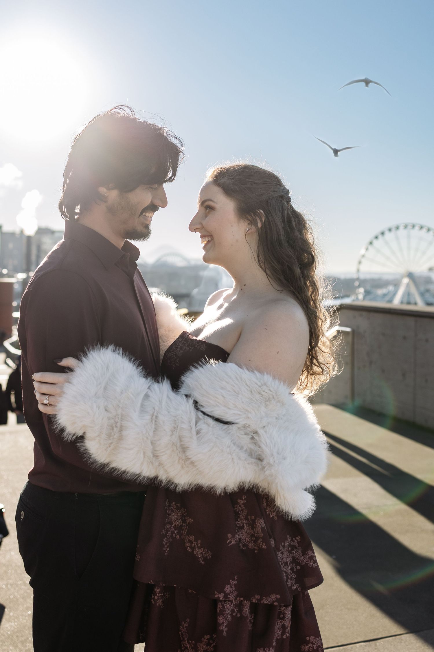 A romantic couple embraces on a rooftop with a Ferris wheel and seagulls visible in the background on a sunny day.