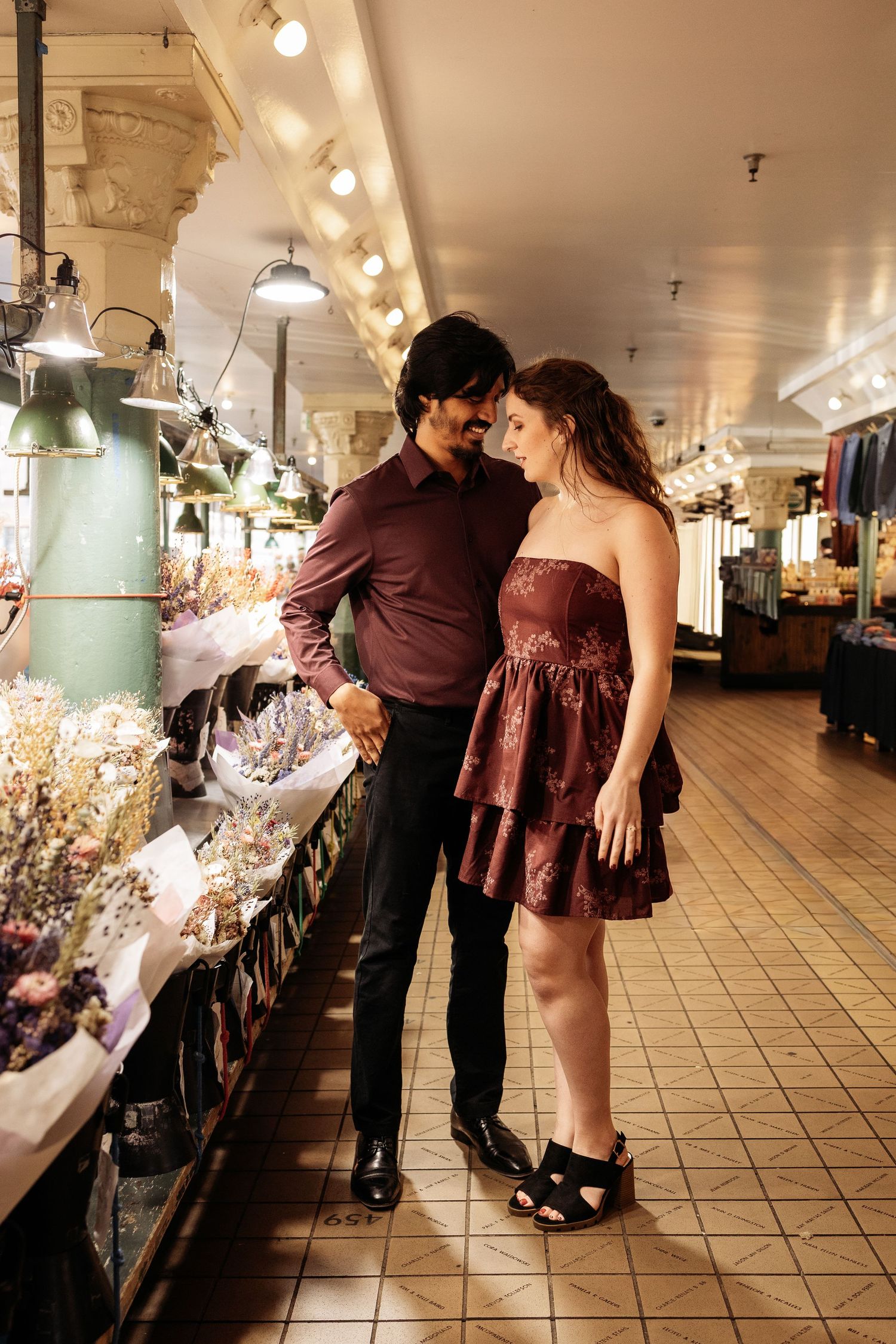 A couple in burgundy outfits dances together in a long tiled hallway with ornate architectural details and lighting.