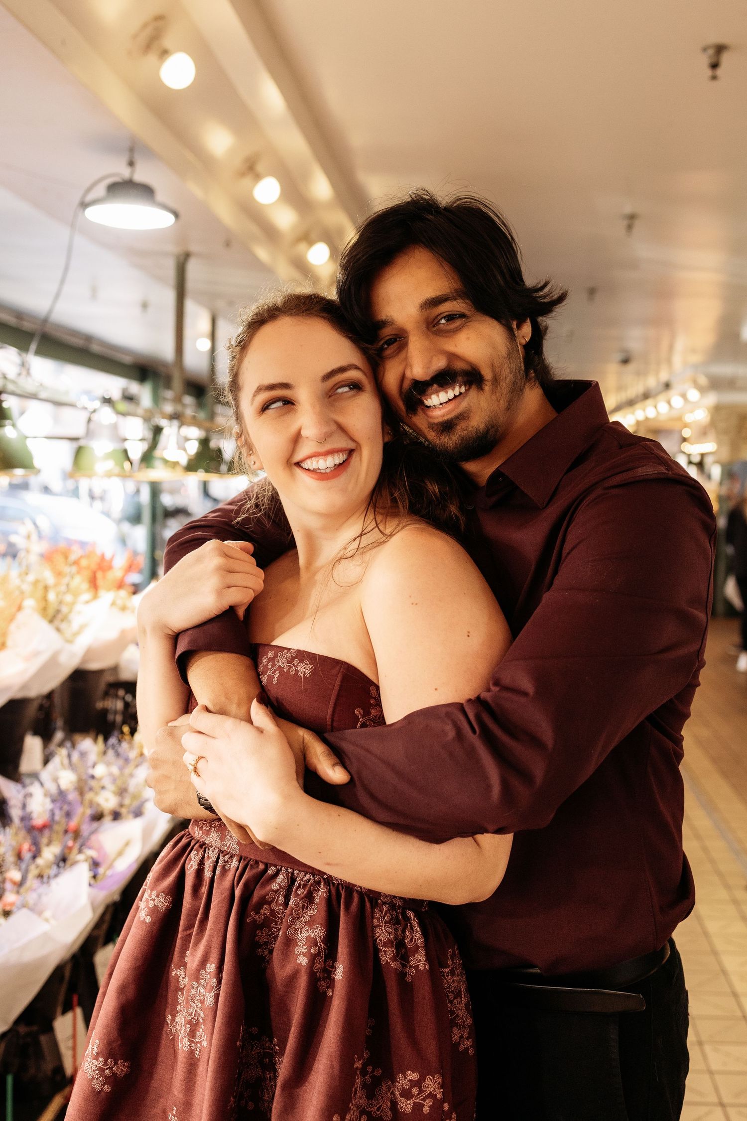 A couple embraces and laughs together in a warmly lit indoor setting wearing burgundy colored outfits.