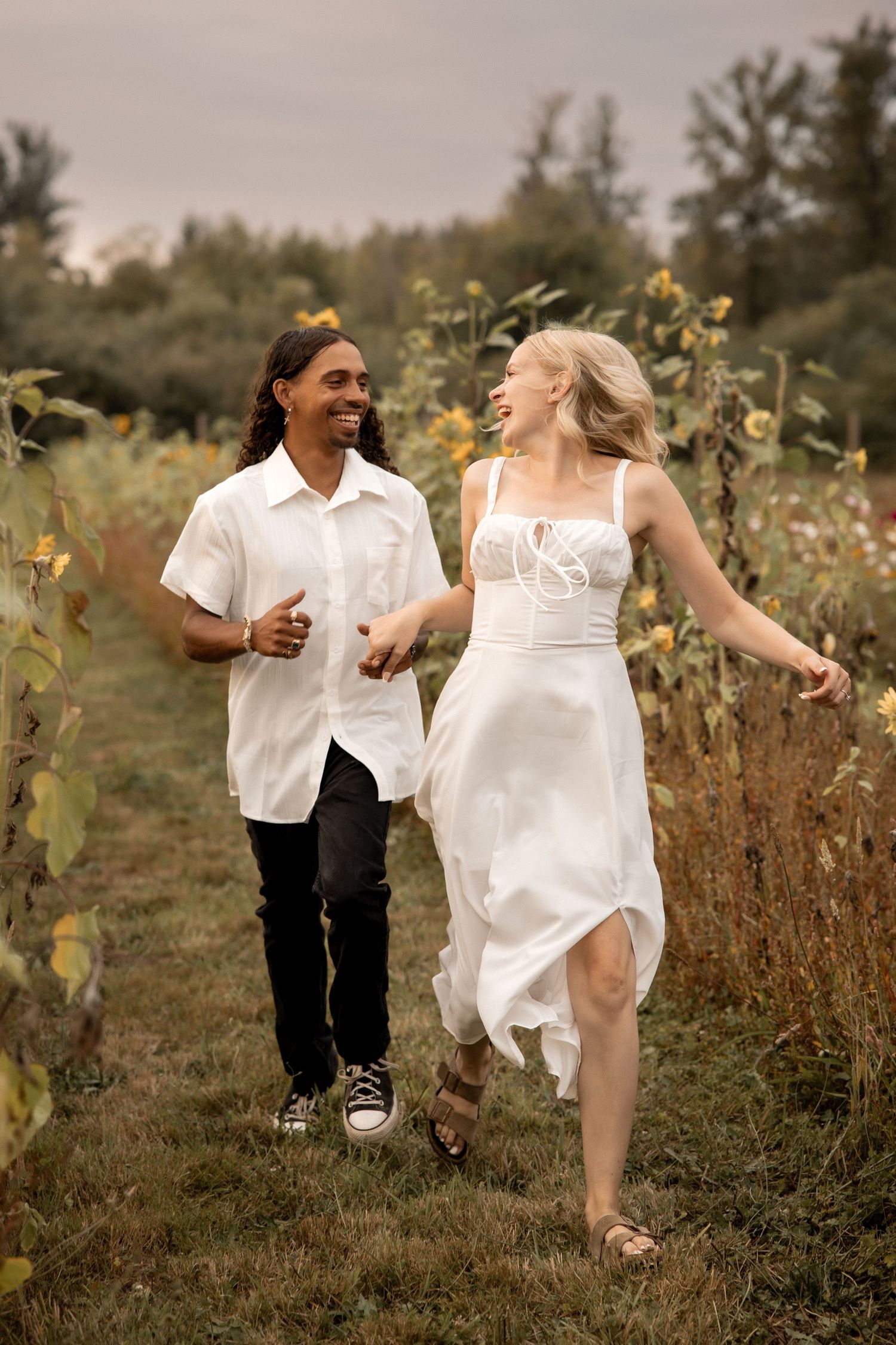 A joyful interracial couple runs through a sunflower field, both smiling, wearing white outfits on a cloudy day.