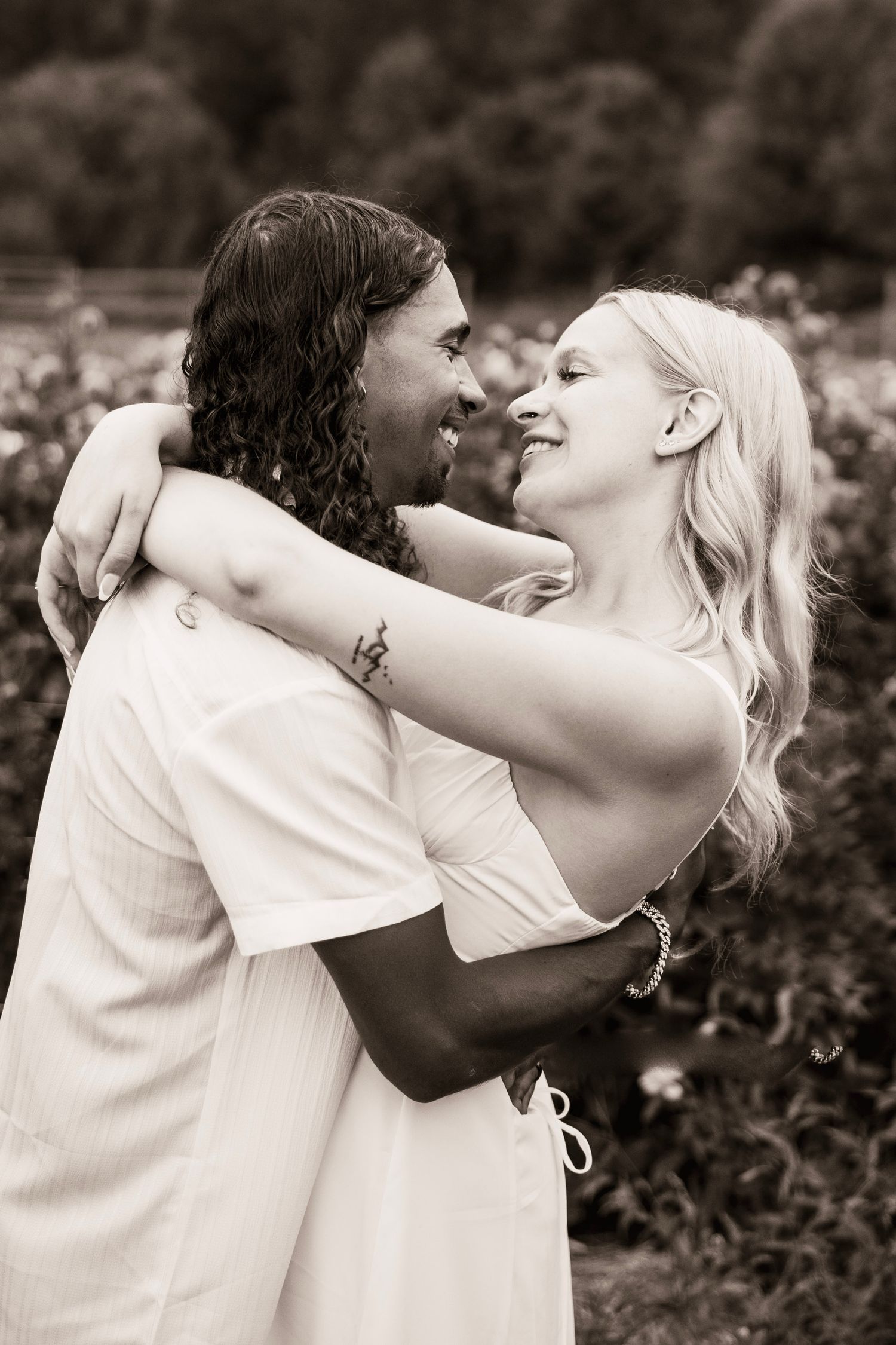 Black and white photo of a happy couple in white outfits embracing and smiling at each other outdoors.