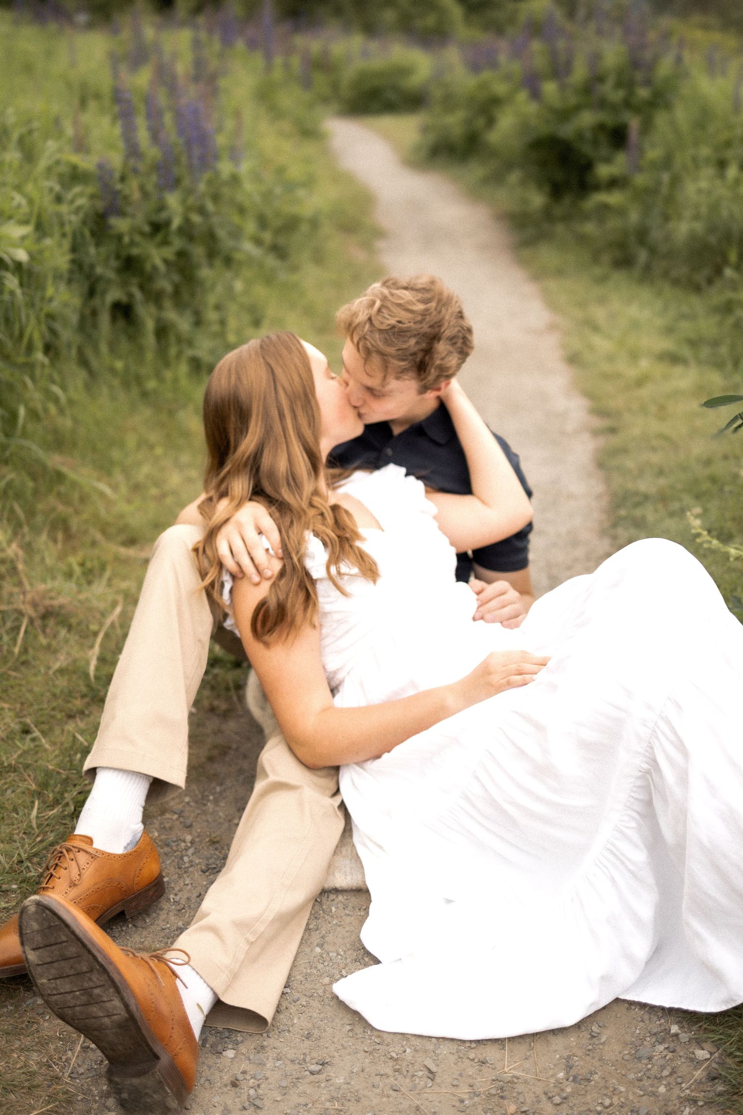 A romantic couple sits together on a dirt path surrounded by summer greenery.