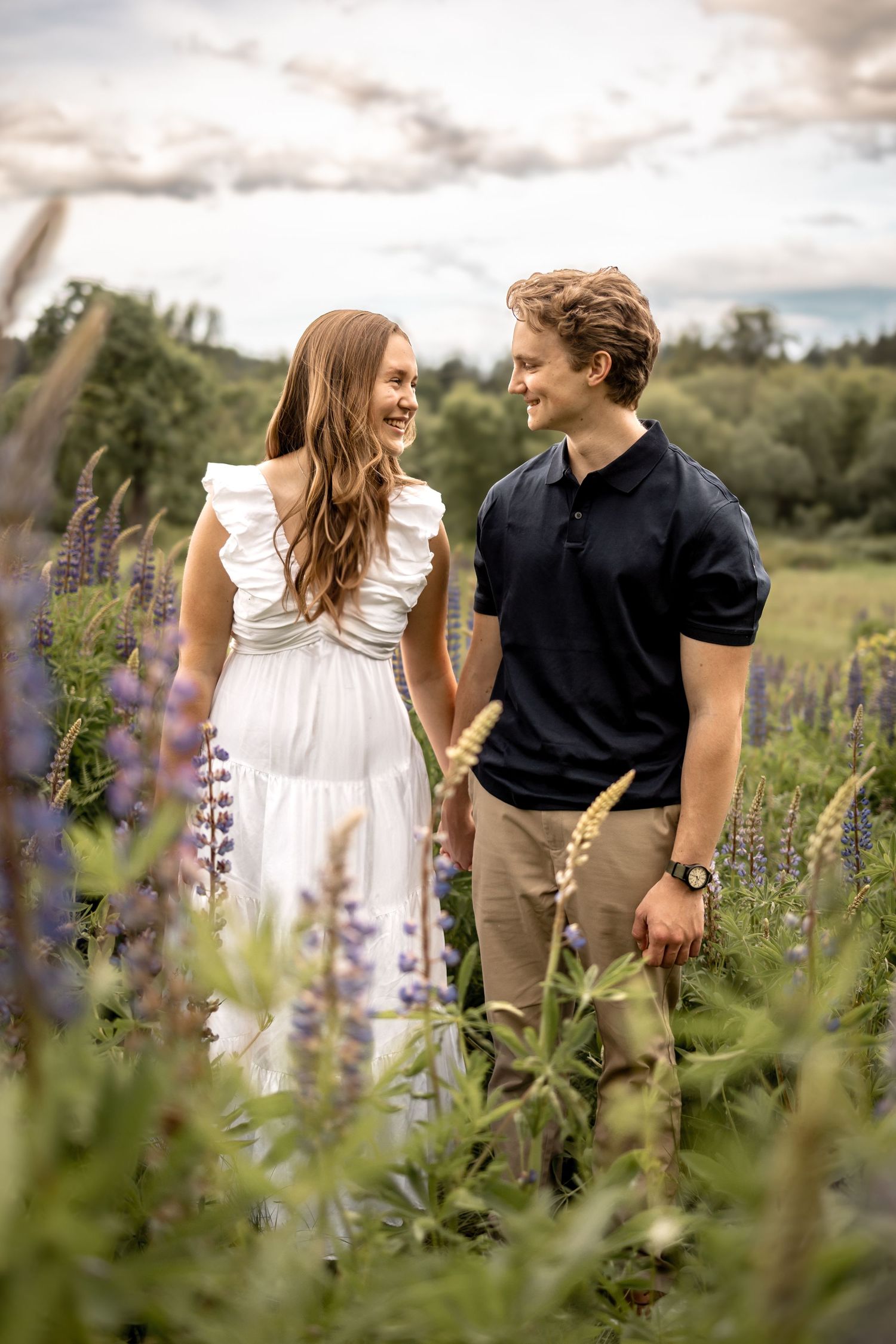 Couple walking hand in hand through field of purple wildflowers at sunset.