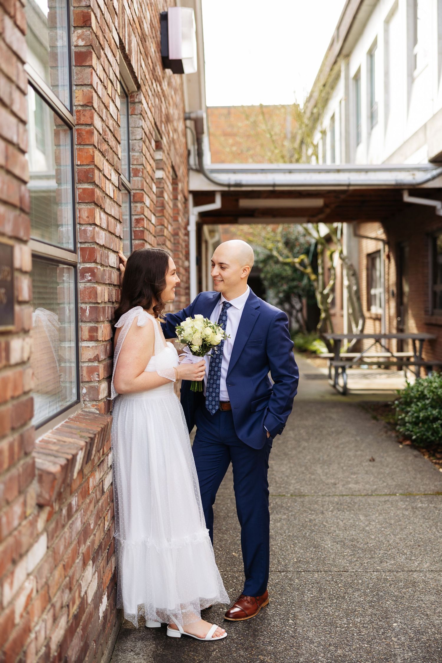 Bride in white dress and groom in navy suit share intimate moments in a brick-lined walkway during wedding photos.