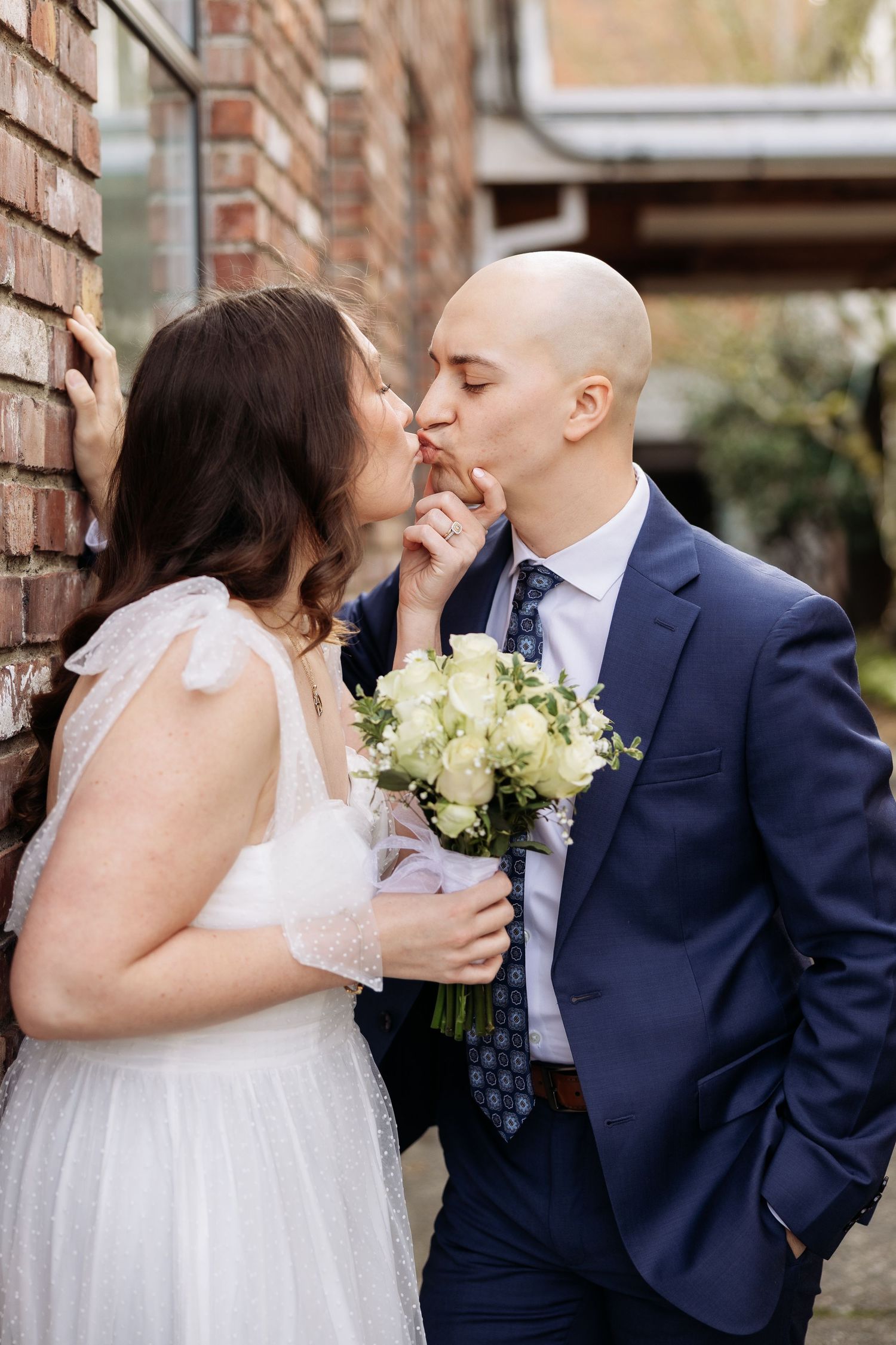A romantic wedding moment against a brick wall as a couple shares a tender kiss while holding a bouquet of white roses.
