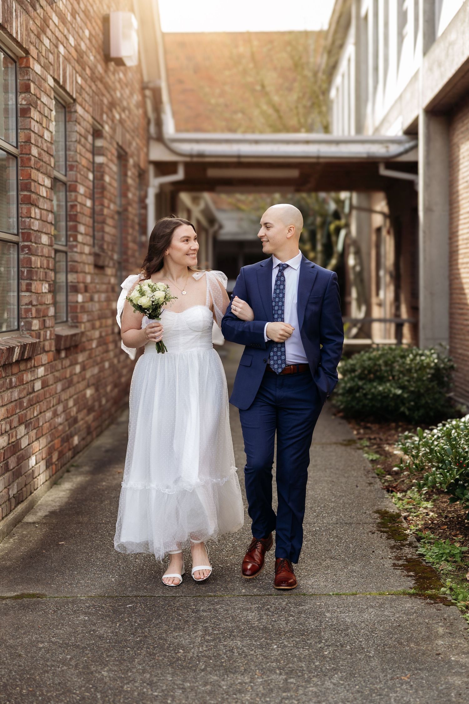 A couple in wedding attire walks down a brick alleyway while holding hands and sharing a sweet moment together.