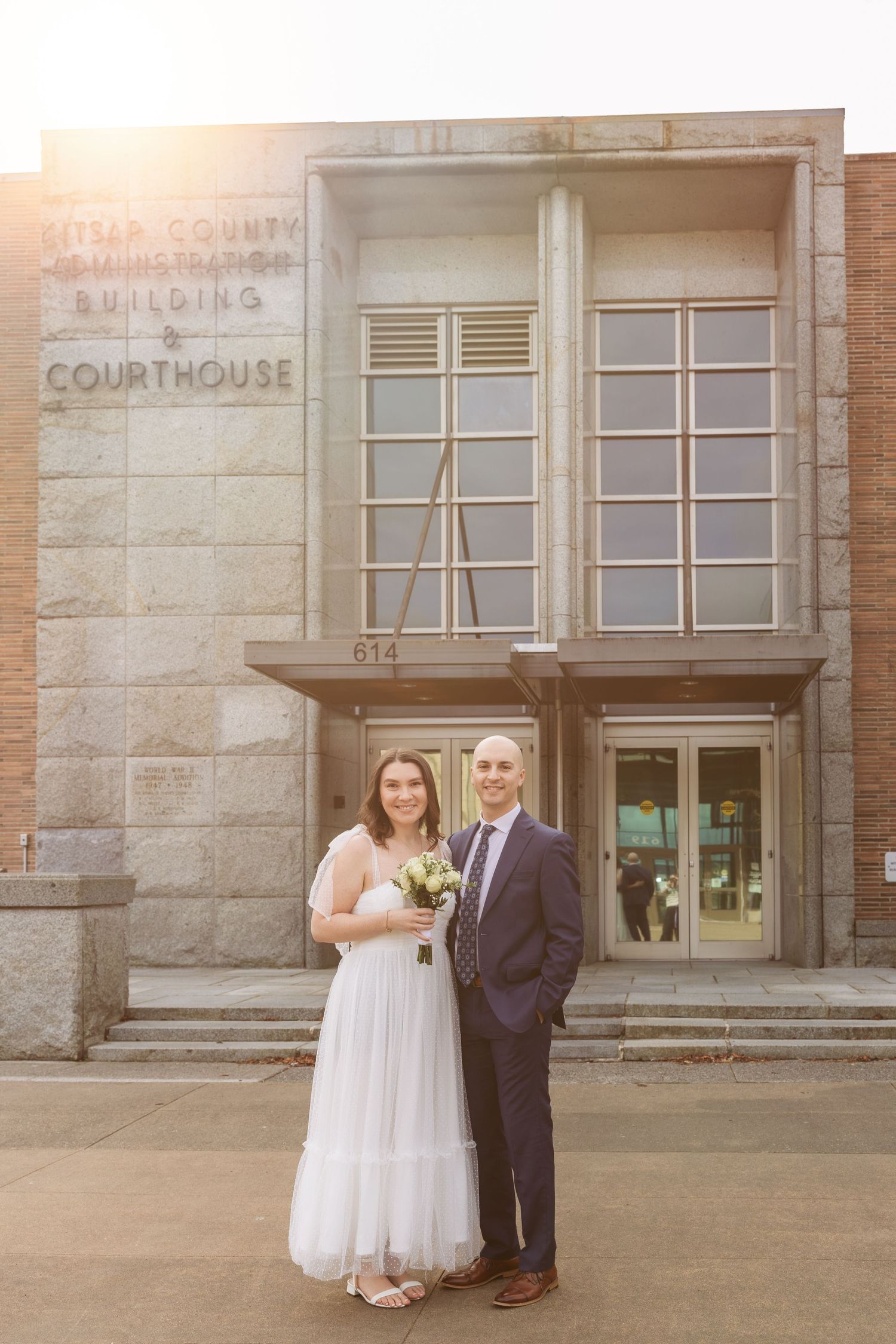 Newlywed couple poses in front of historic courthouse building with stone facade and windows in warm sunlight.