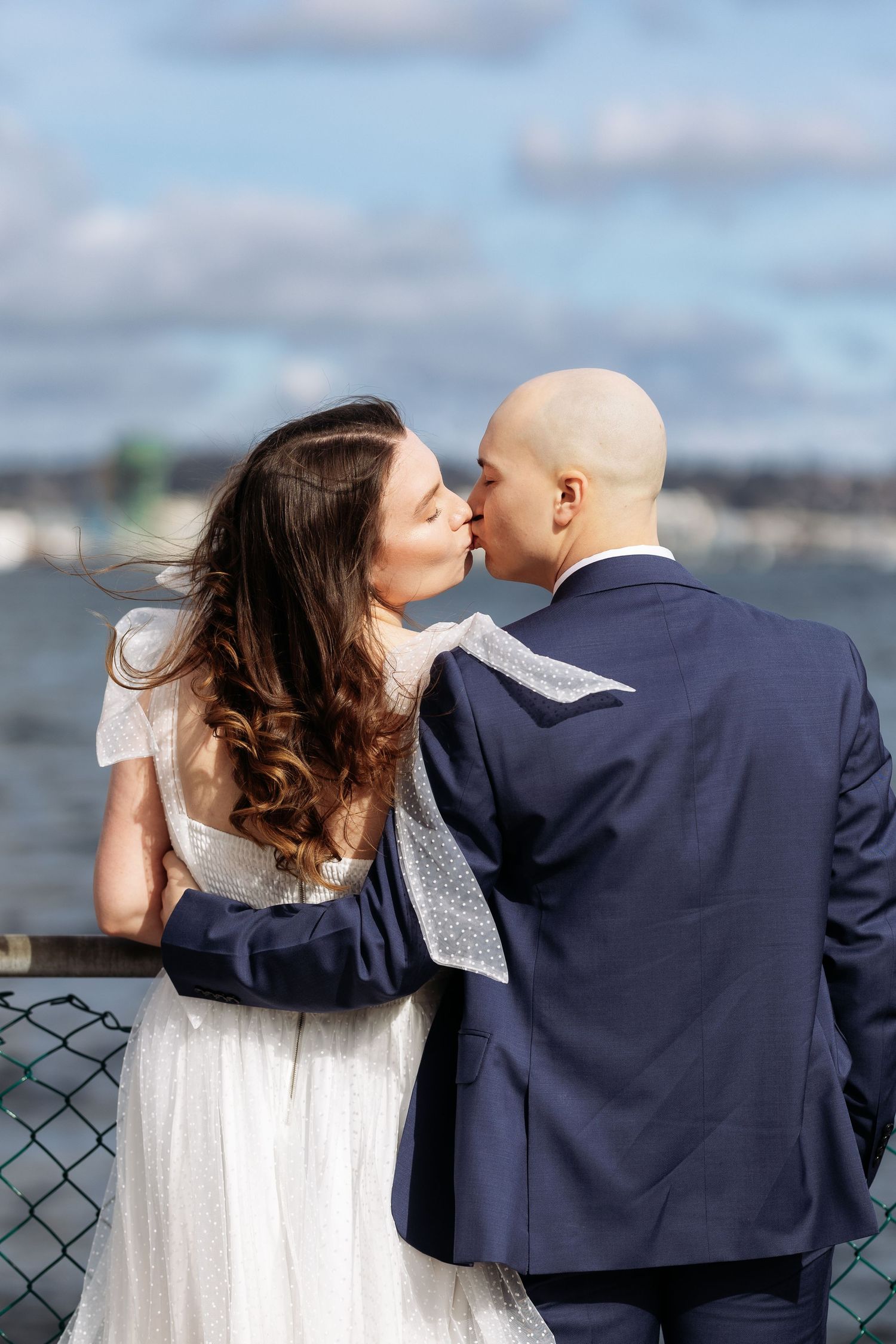 A romantic couple shares a tender kiss while leaning against a railing with a waterfront view in the background.