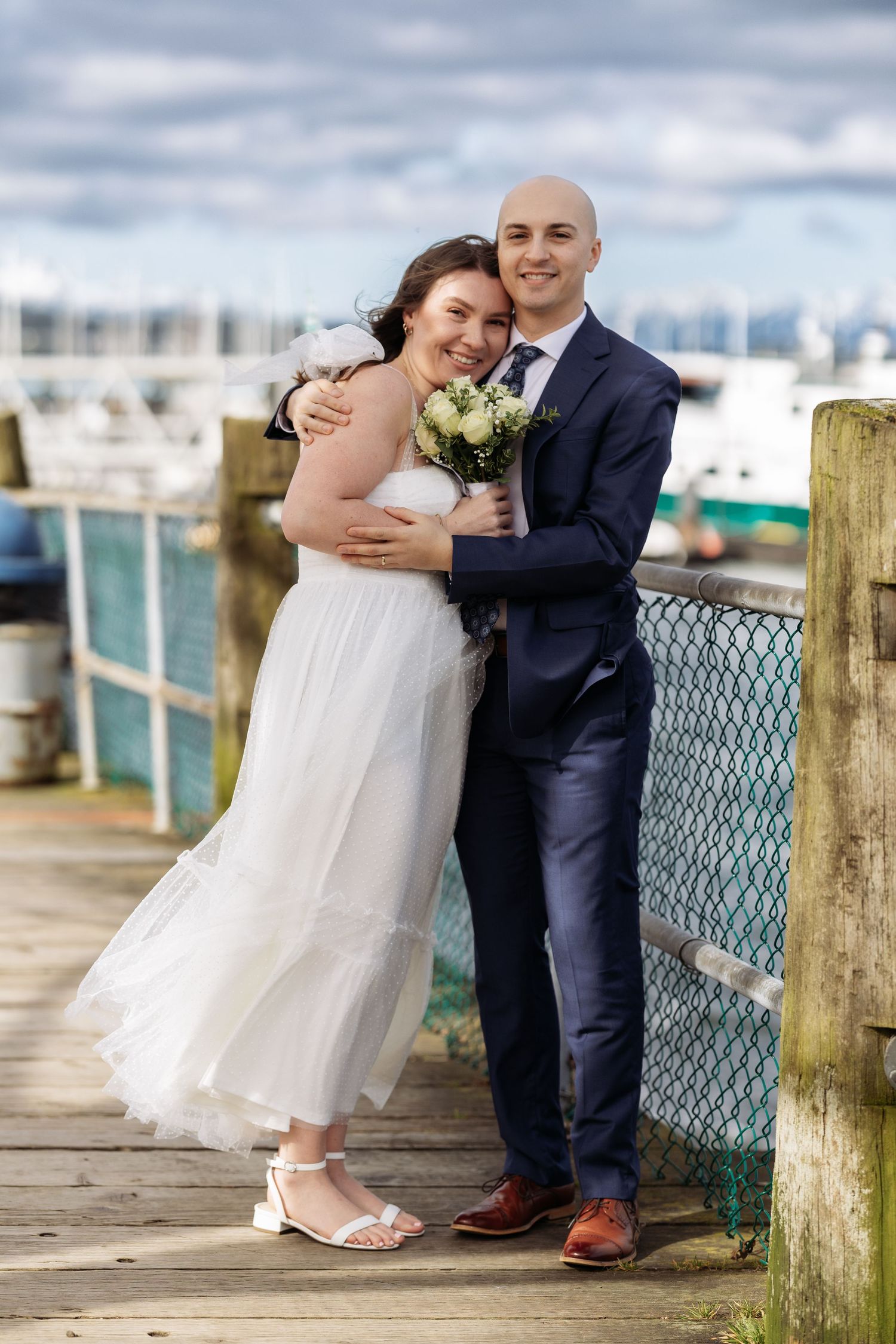 A couple in wedding attire shares romantic moments along a waterfront boardwalk with wooden posts and blue railing.