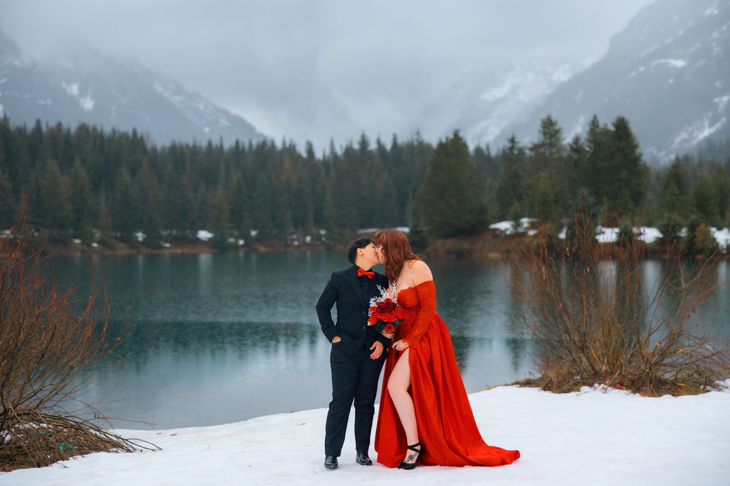 A couple in winter attire stands near a snow-covered lake with mountains and evergreen forest in the background.