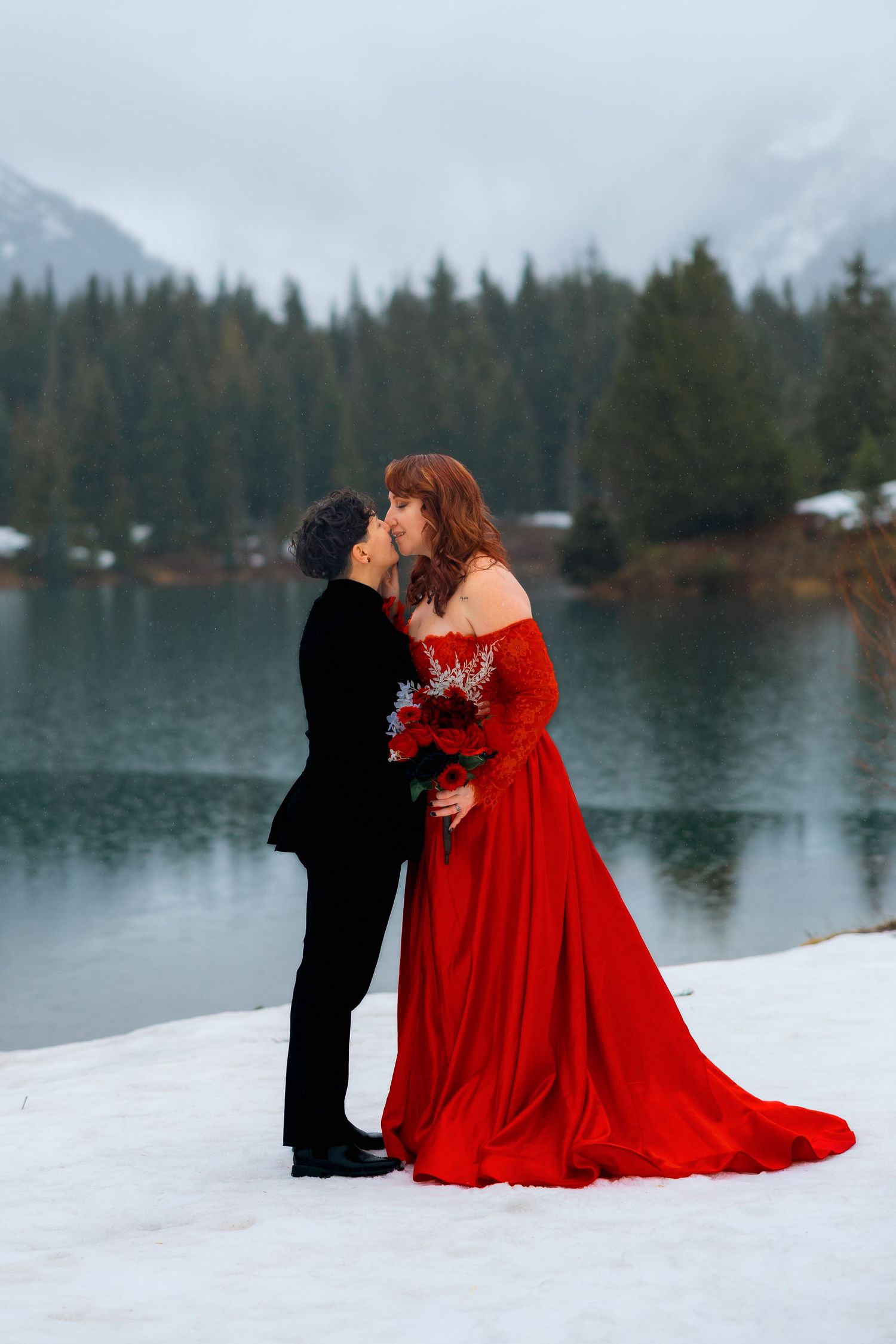 A couple in formal attire shares a romantic moment by a snowy lake with evergreen trees in the background.