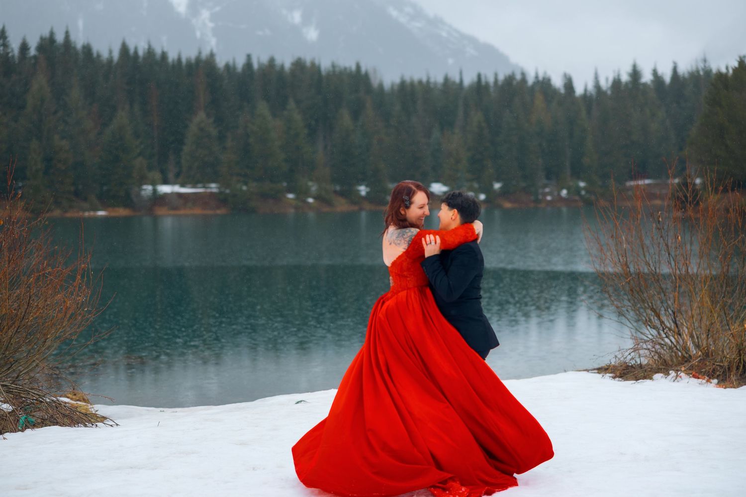 A couple embraces by a serene mountain lake in winter, with the person in a flowing red dress against snowy ground.