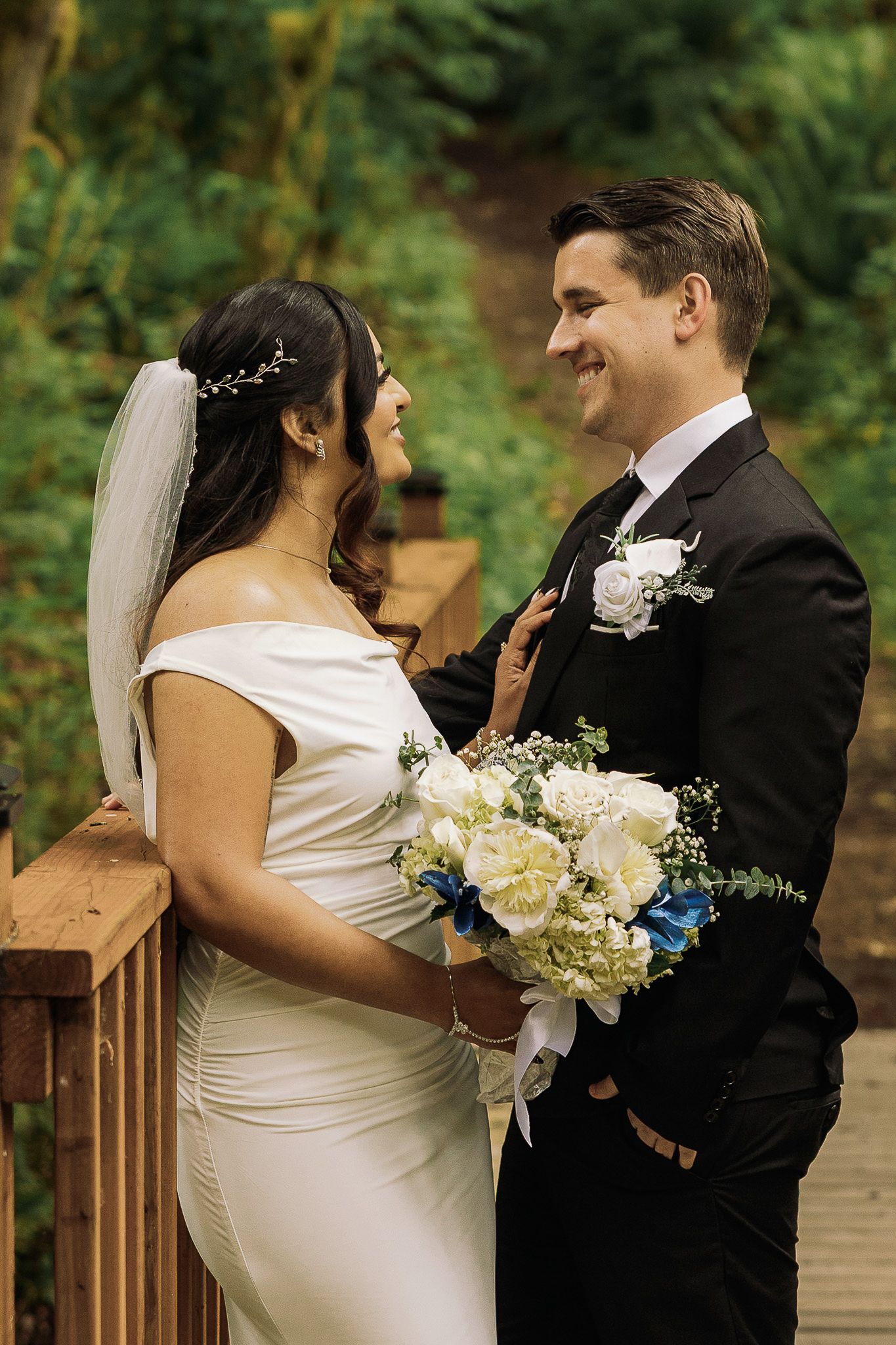 Bride and groom smiling at each other on a wooden deck, bride holding white and blue bouquet, lush green forest background.