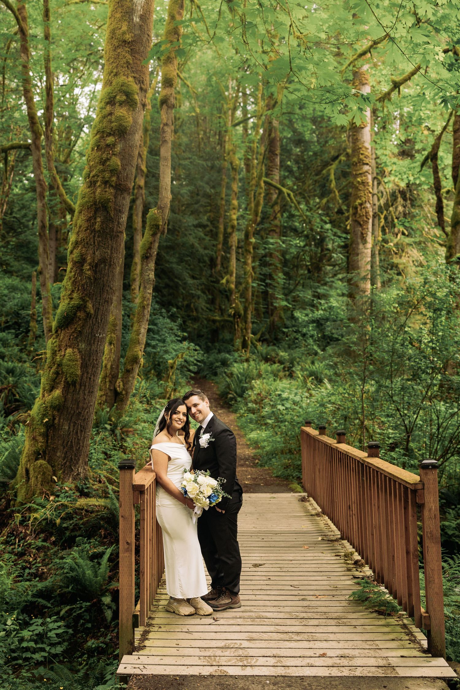 Bride and groom pose on a wooden bridge surrounded by lush, moss-covered trees in a forest wedding setting.