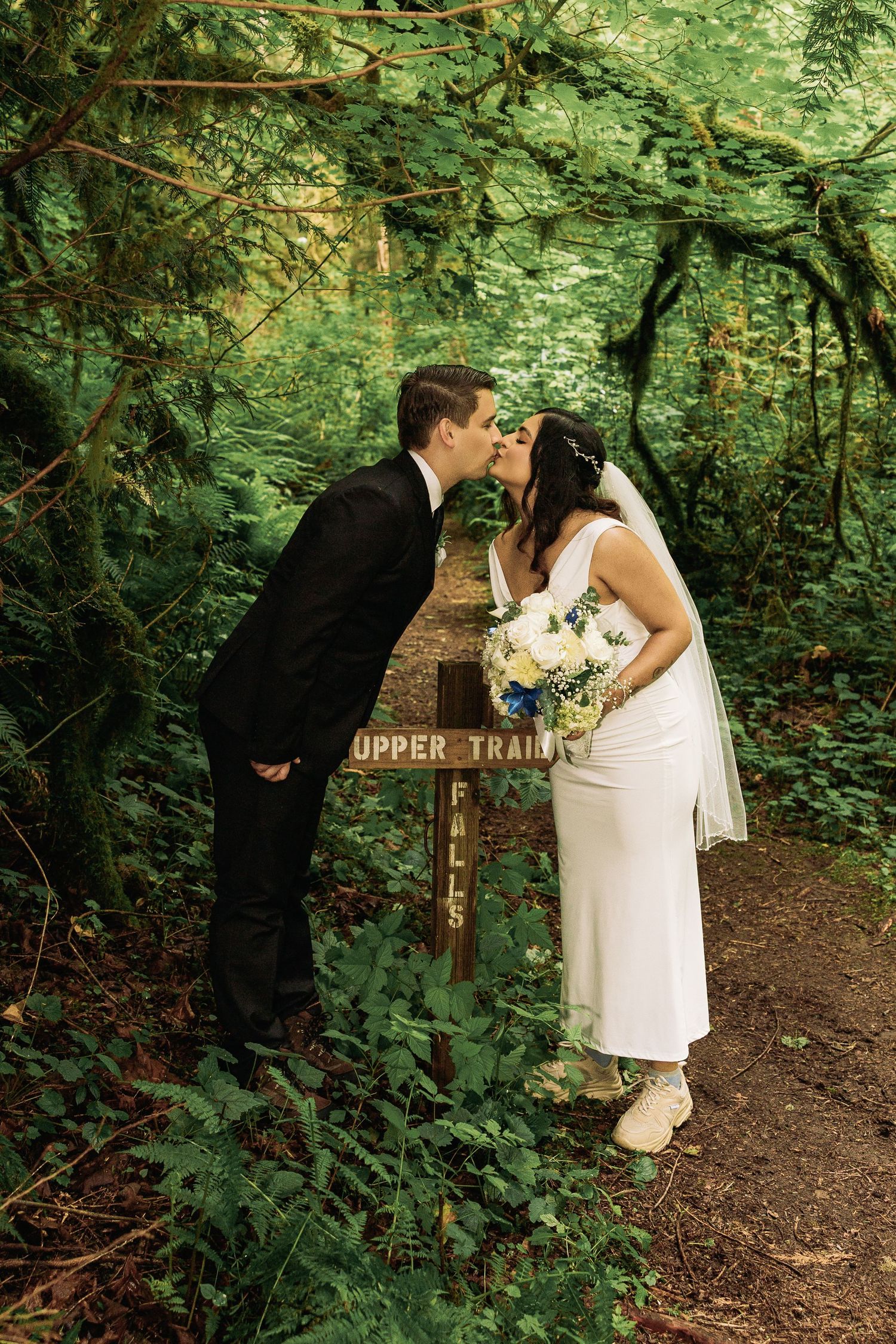 Bride and groom kissing beside Upper Trail Falls sign surrounded by lush green forest, bride holding white floral bouquet.