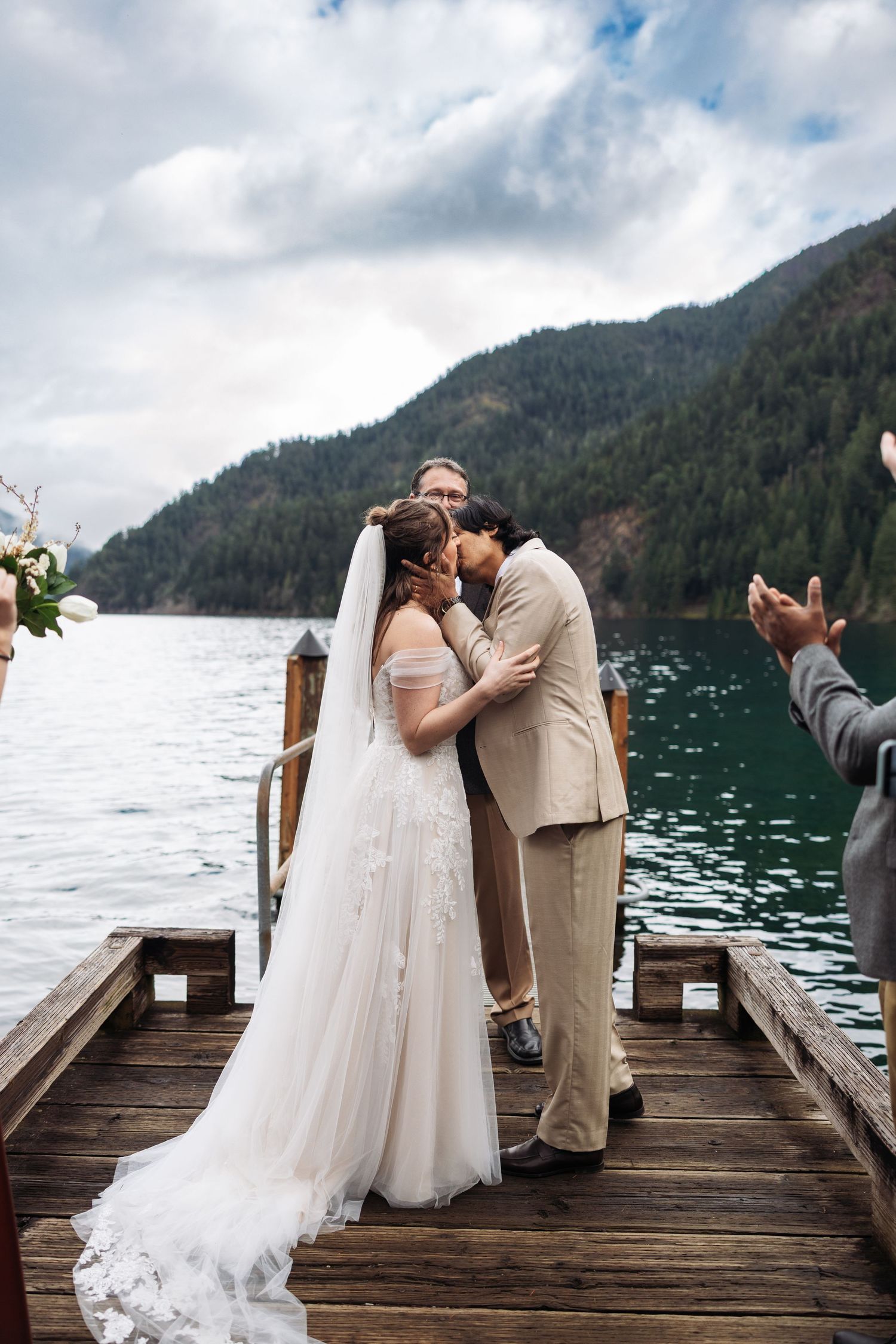 A couple shares an intimate moment on a wooden dock overlooking a scenic mountain lake surrounded by evergreen forests.