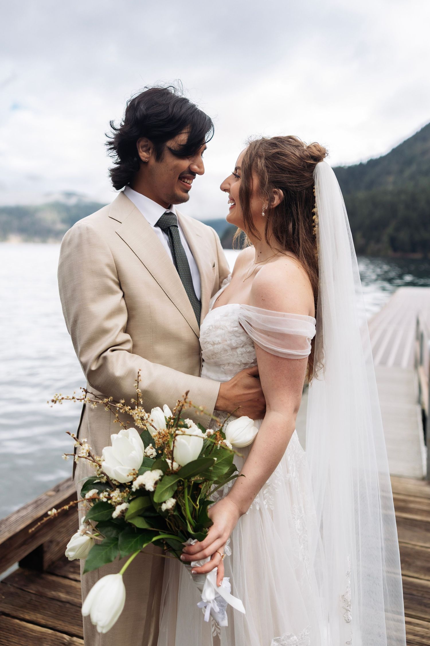 Newlywed couple shares intimate moment on wooden dock by lake with mountains and cloudy sky in background.