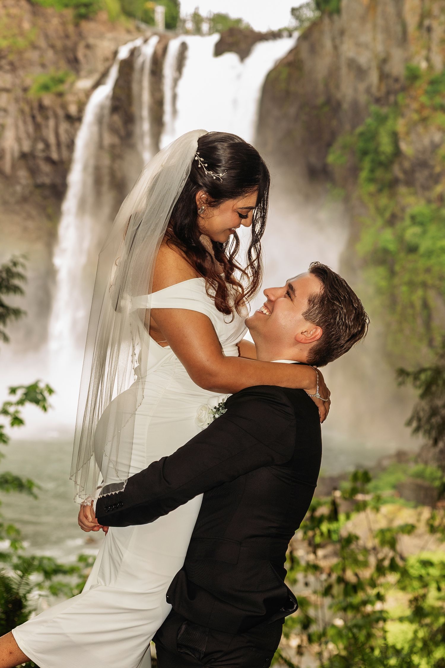 Groom lifts bride in white gown before stunning waterfall backdrop in lush green outdoor wedding photo.