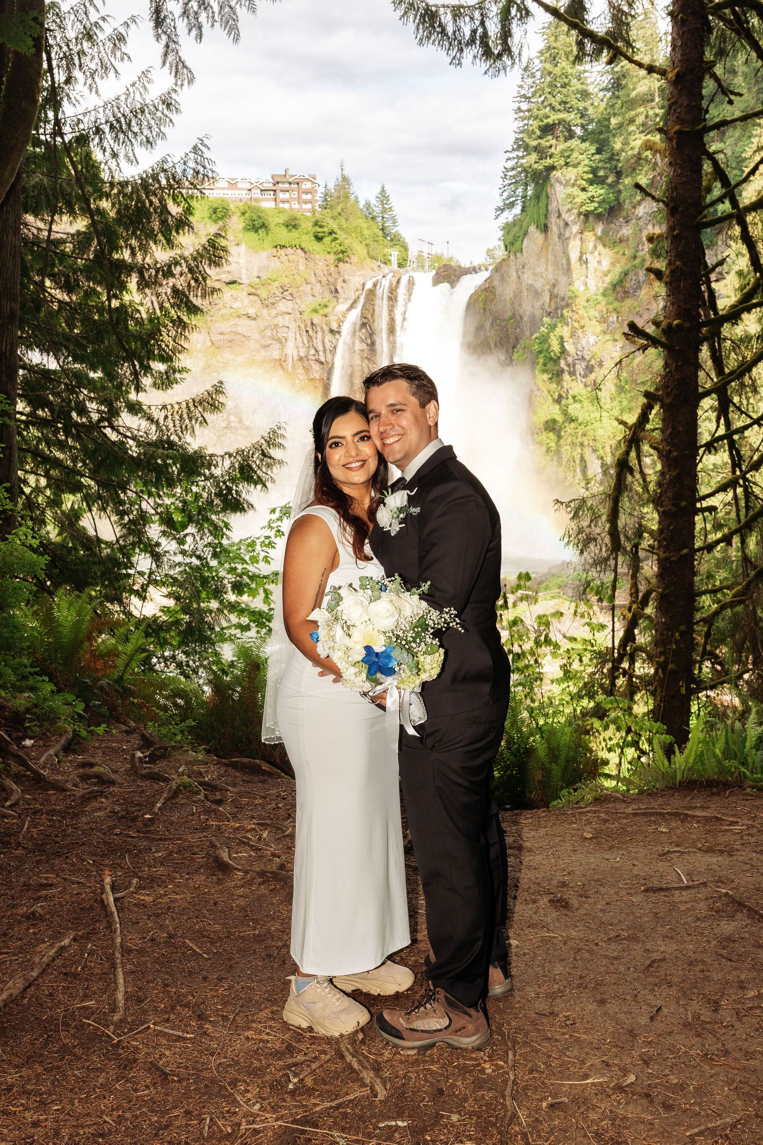 Bride and groom pose in front of Snoqualmie Falls waterfall, bride holding white and blue bouquet in lush forest setting.
