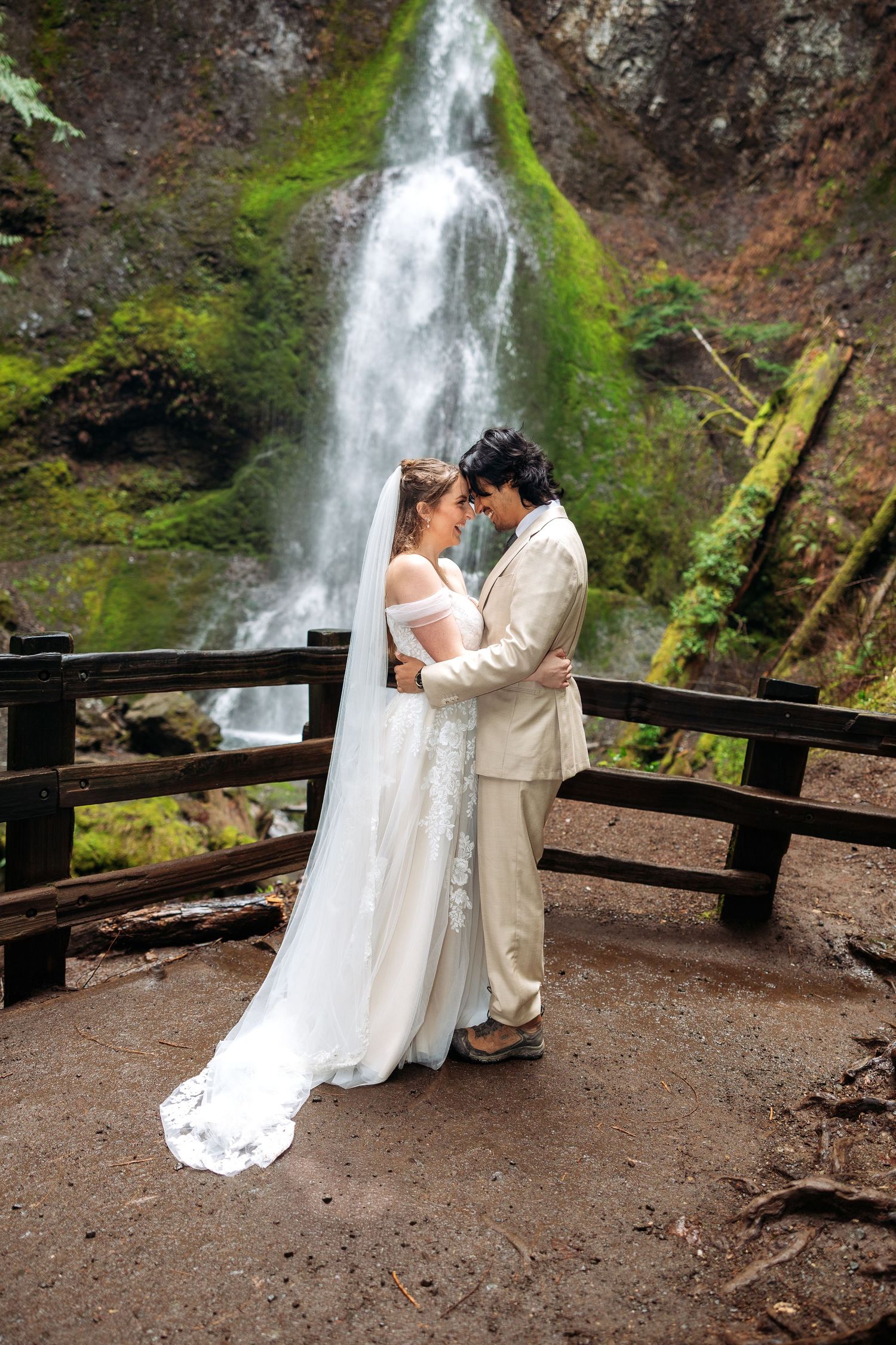 Bride and groom embrace before a lush waterfall on a wooden bridge, bride in white gown with veil, groom in tan suit.