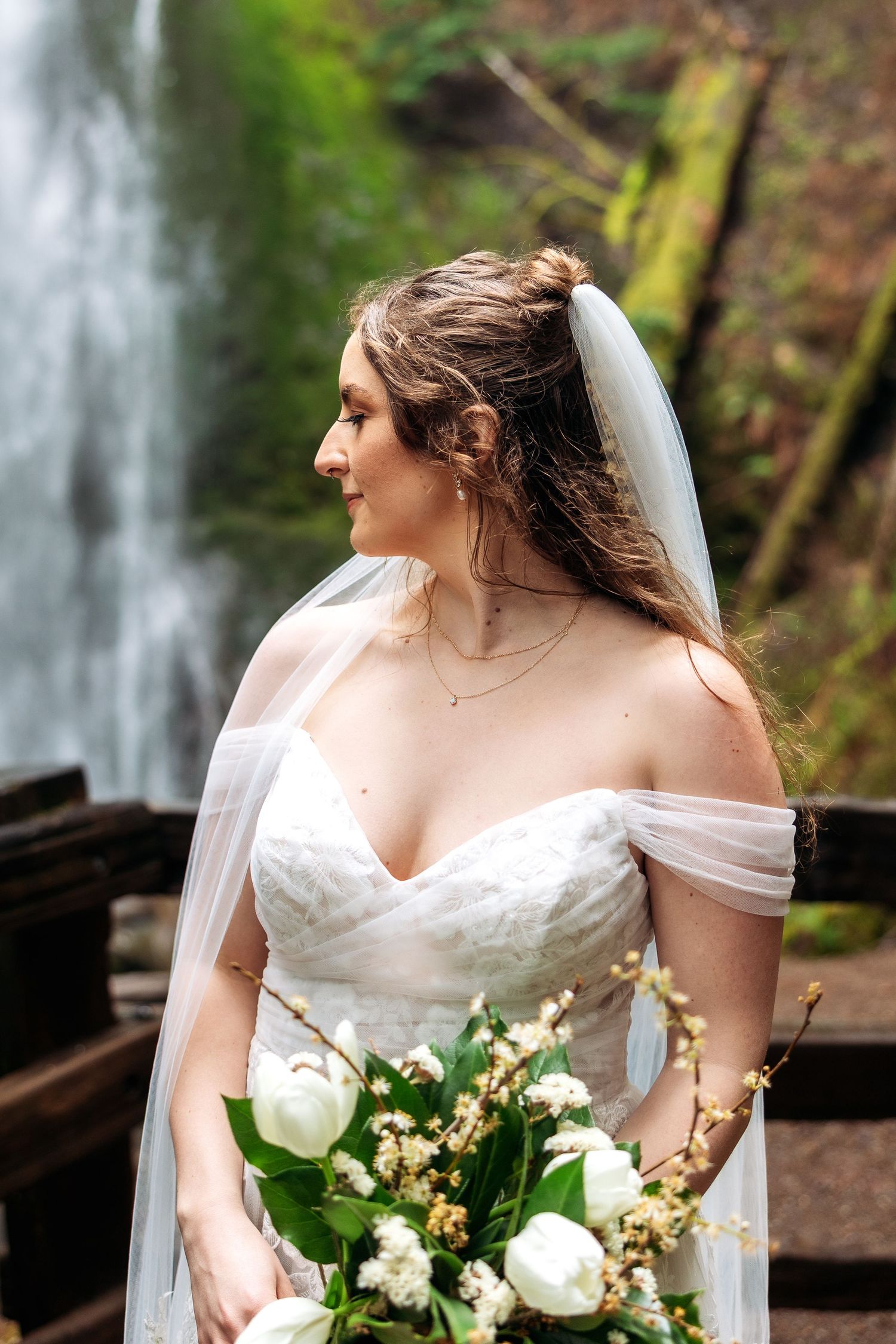 Bride in white lace off-shoulder gown holds white tulip bouquet before a lush waterfall backdrop.
