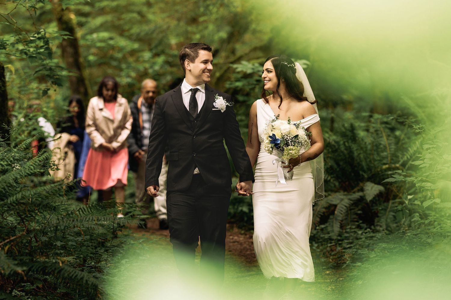 Bride and groom walk hand-in-hand through a lush green forest during an outdoor wedding ceremony.