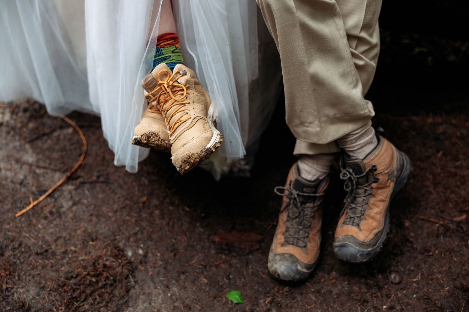 Two pairs of hiking boots standing on muddy ground with a white dress and khaki pants visible above them.