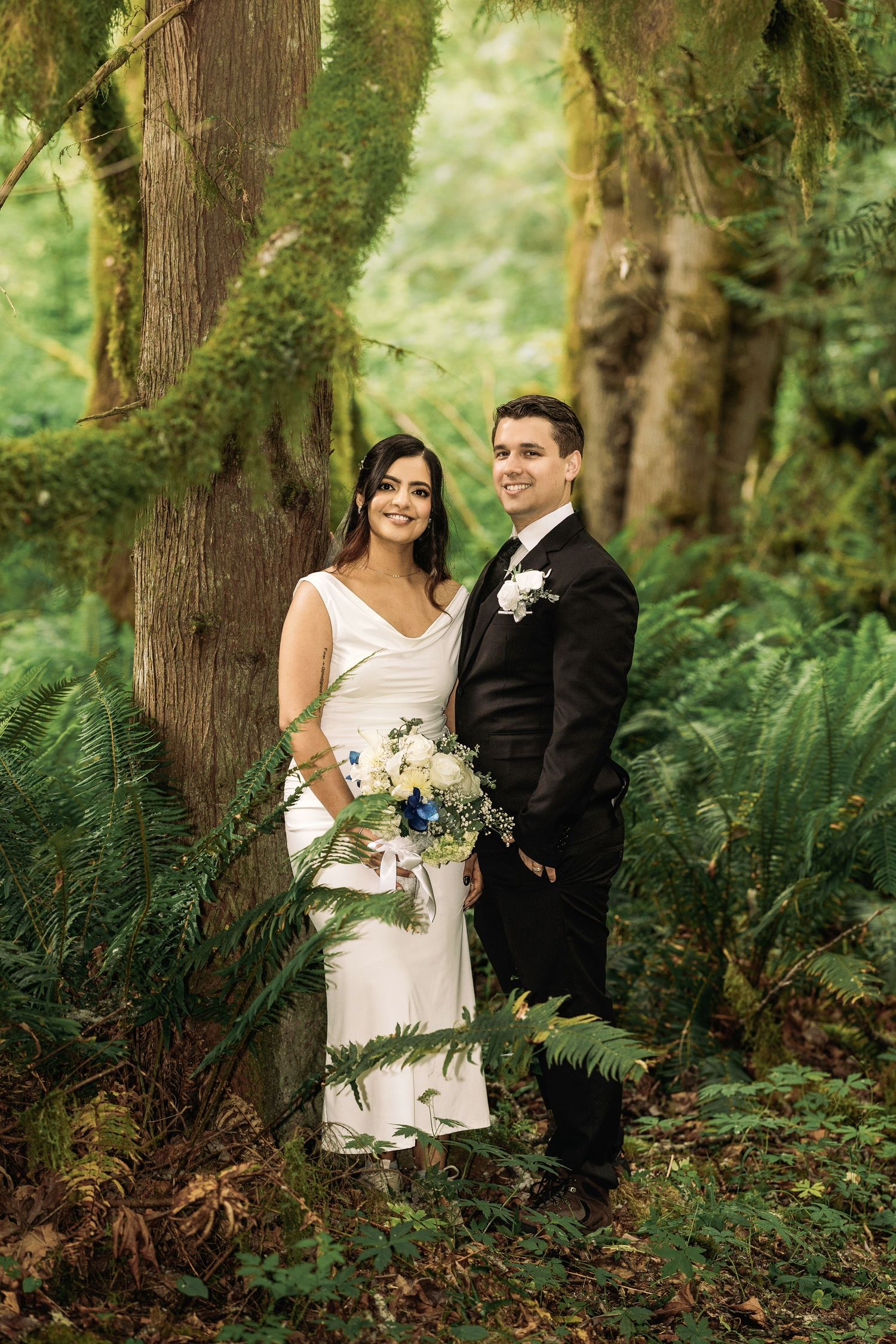 Bride and groom pose in mossy Pacific Northwest forest, bride holding white and blue bouquet.