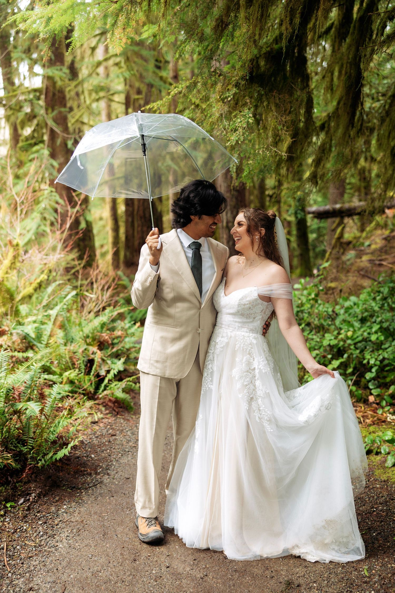 A romantic wedding couple walks along a mossy forest path under a white umbrella in the Pacific Northwest.