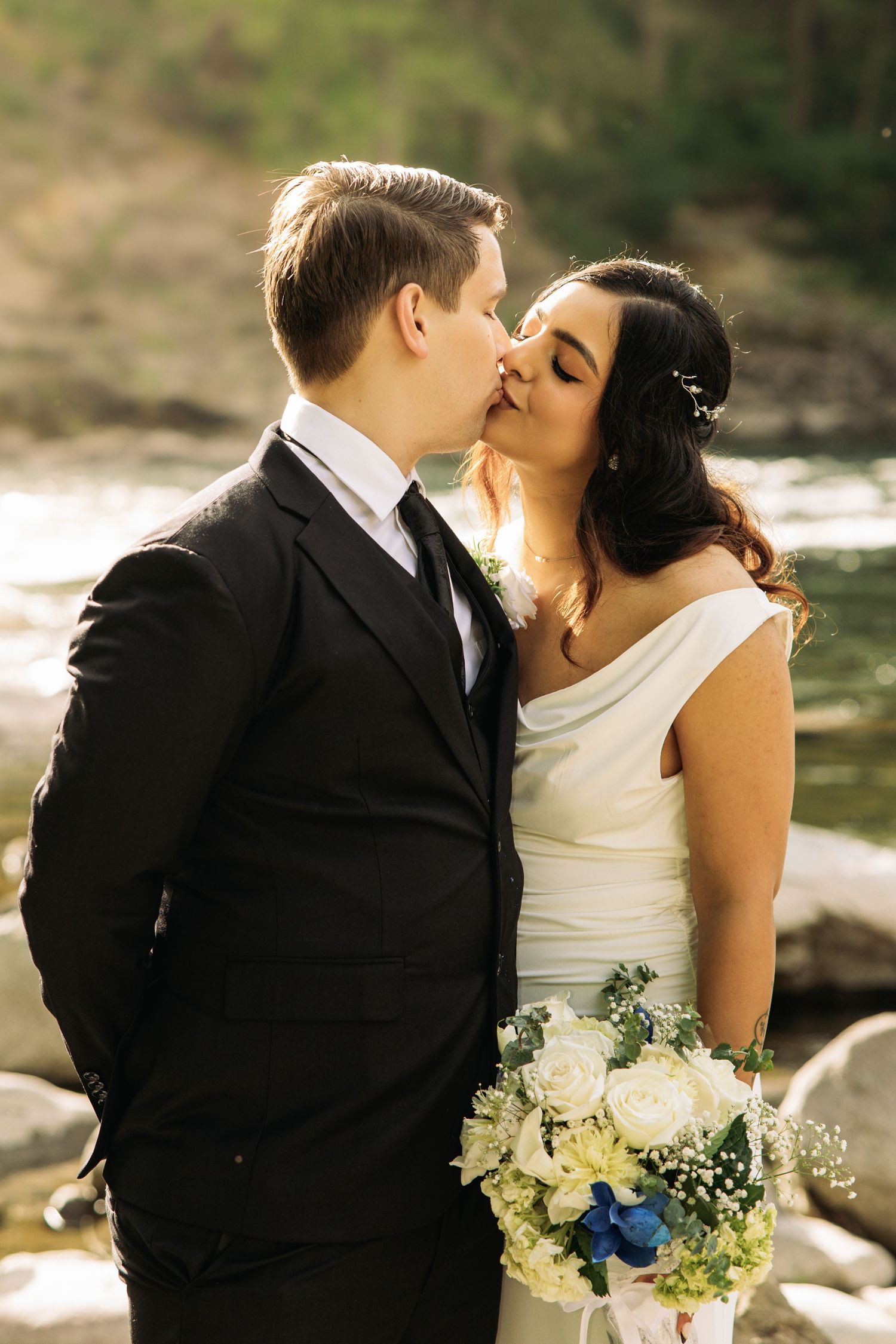 Bride and groom share a romantic kiss outdoors by a river, bride holding a white and blue bouquet.