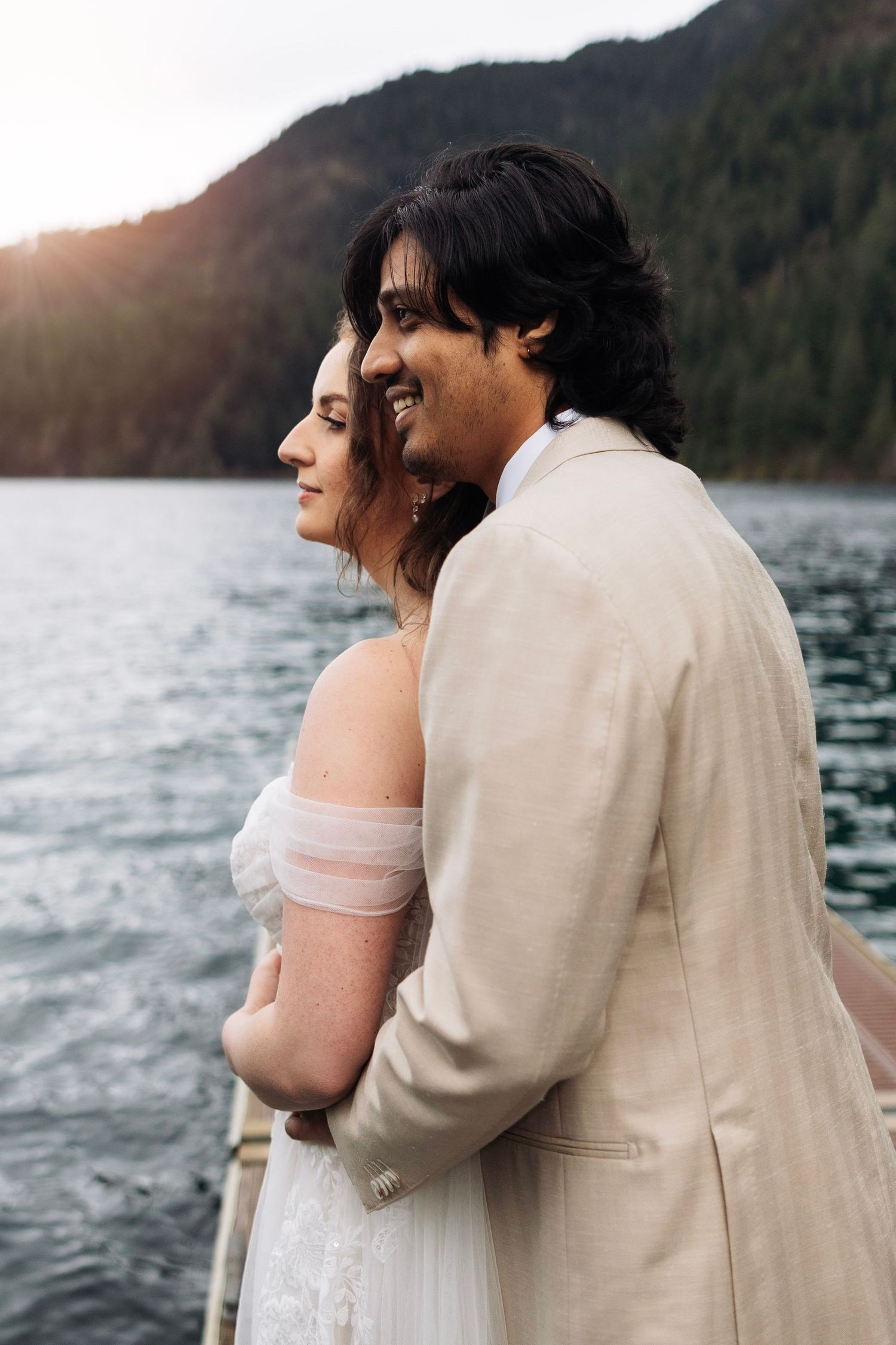 A couple in wedding attire embraces by a scenic lake at sunset with mountains in the background.