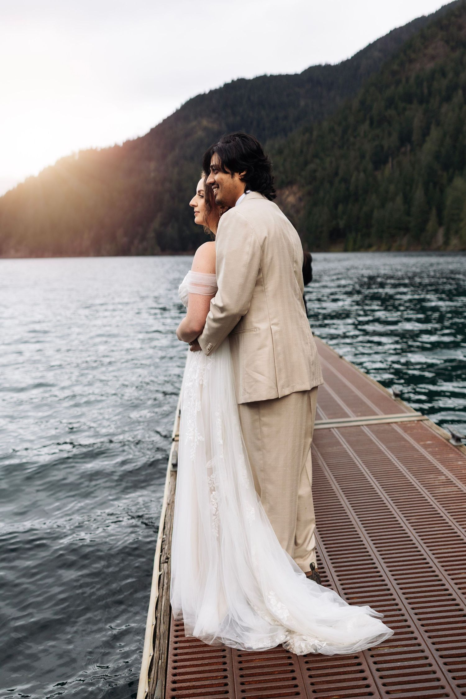 Newlyweds share an intimate moment on a wooden dock overlooking a misty mountain lake at sunset.