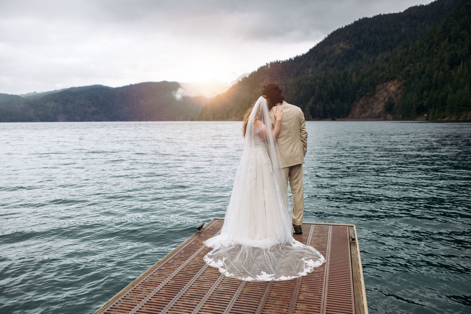 Scenic lakeside dock wedding ceremony with mountain backdrop and dramatic sunset over water.