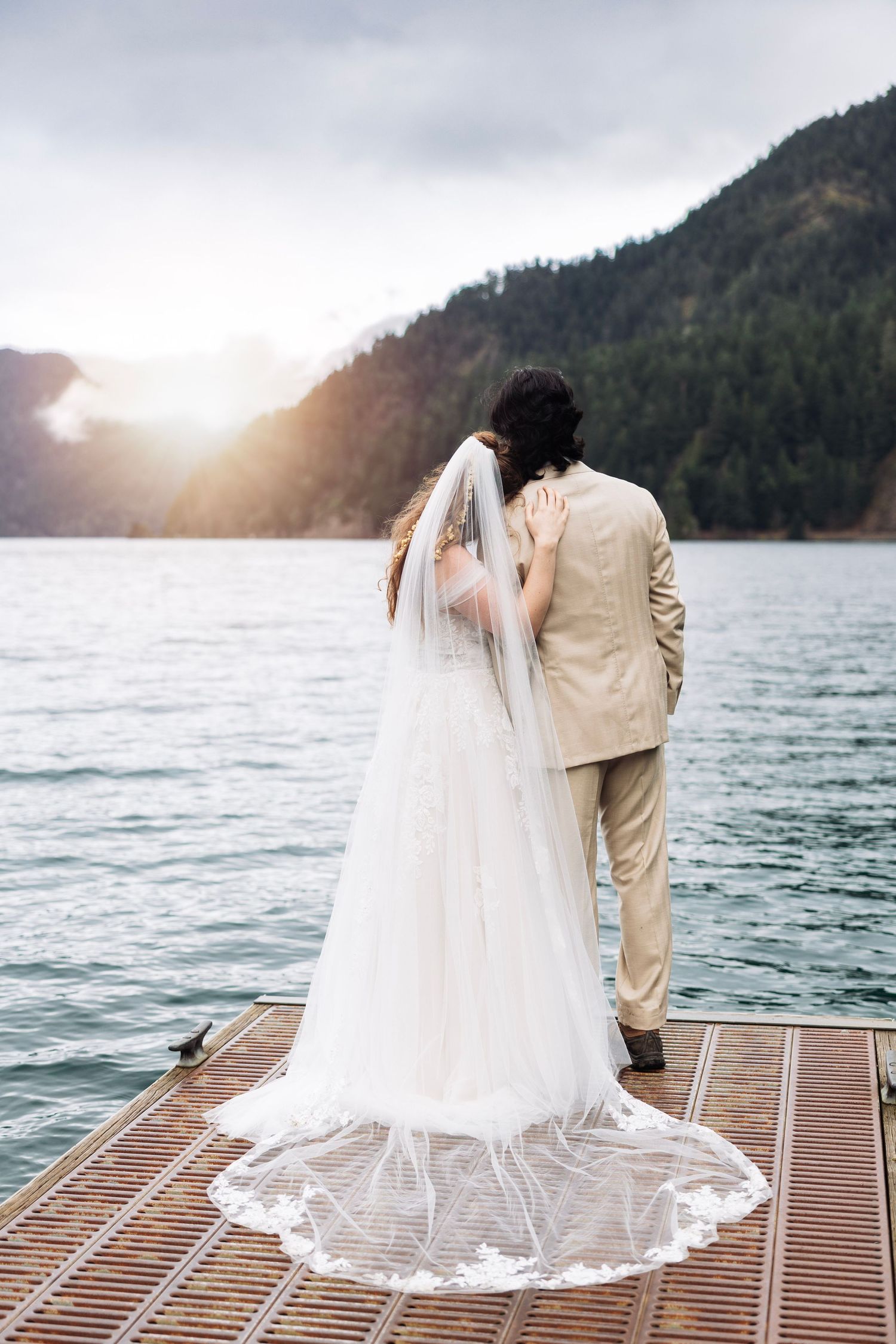 Newlyweds sit together on a wooden dock overlooking a scenic mountain lake at sunset.