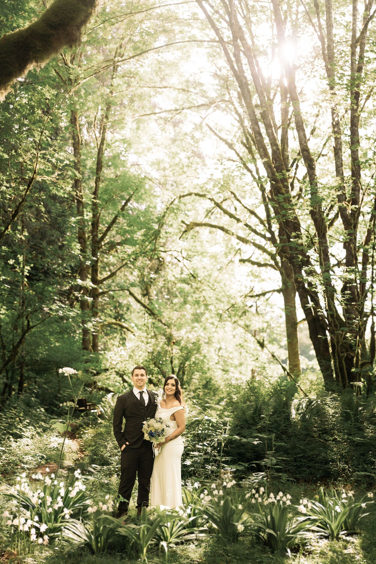 Bride and groom standing together in a sunlit forest, surrounded by lush greenery and white flowers.