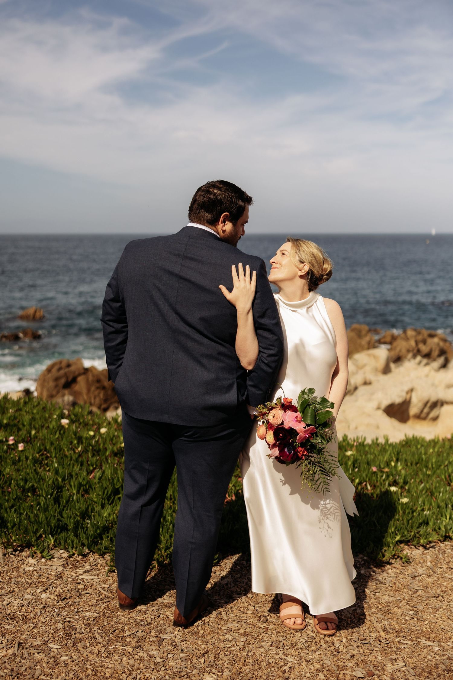 A couple shares a joyful moment on a coastal cliff overlooking the ocean, with the bride holding a vibrant bouquet.