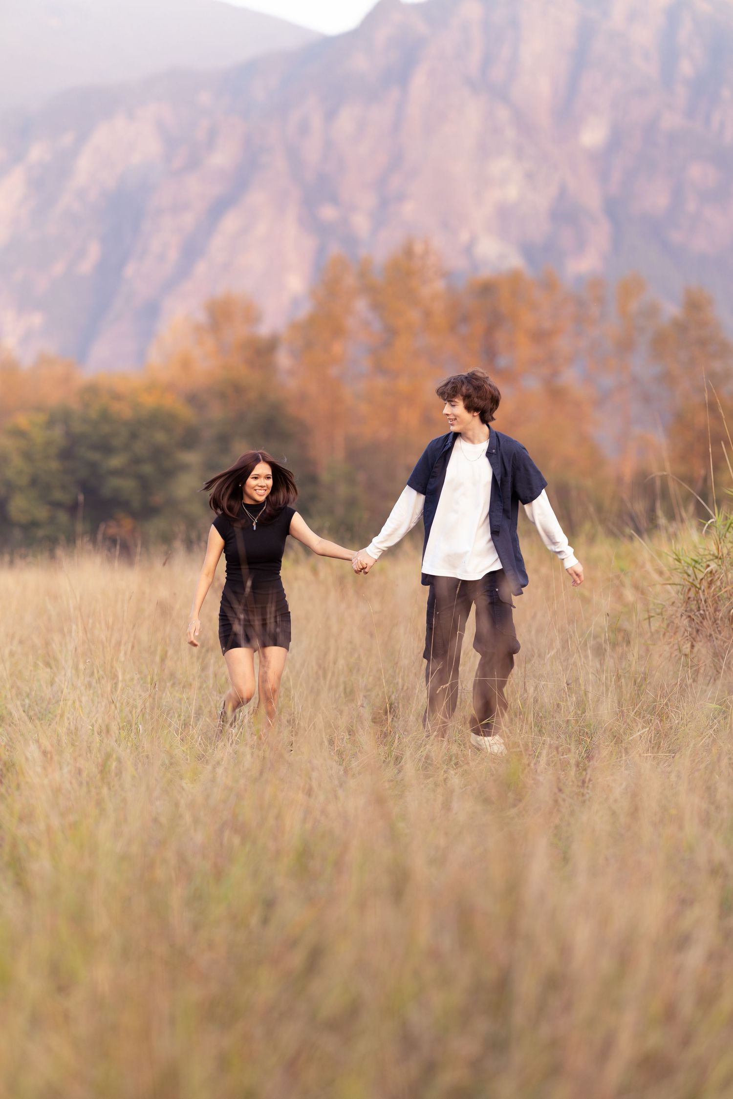 Couple playfully running and dancing together in a golden field with mountains in the background.