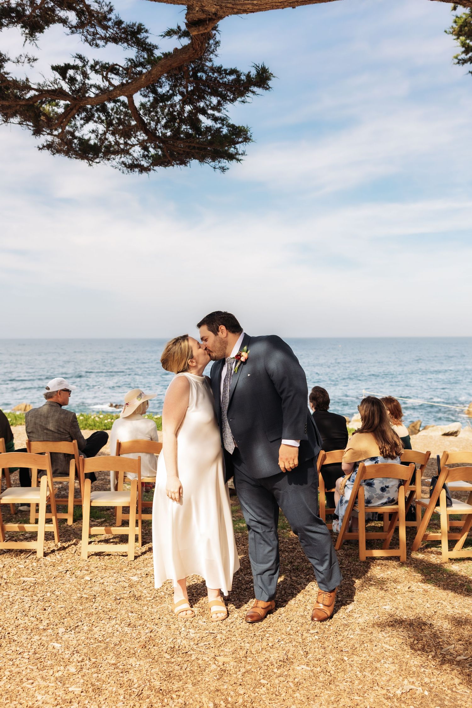 A romantic outdoor wedding ceremony takes place on a cliff overlooking the ocean with wooden chairs and a tree overhead.