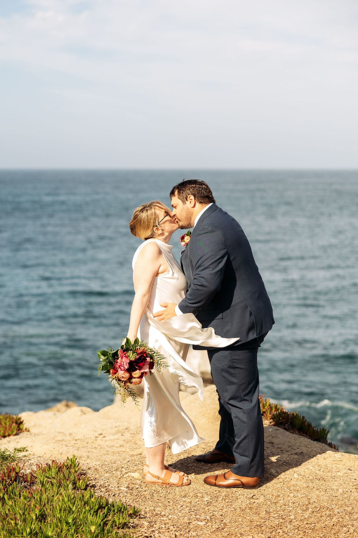 A romantic wedding couple shares a kiss on a coastal cliff overlooking the blue ocean.