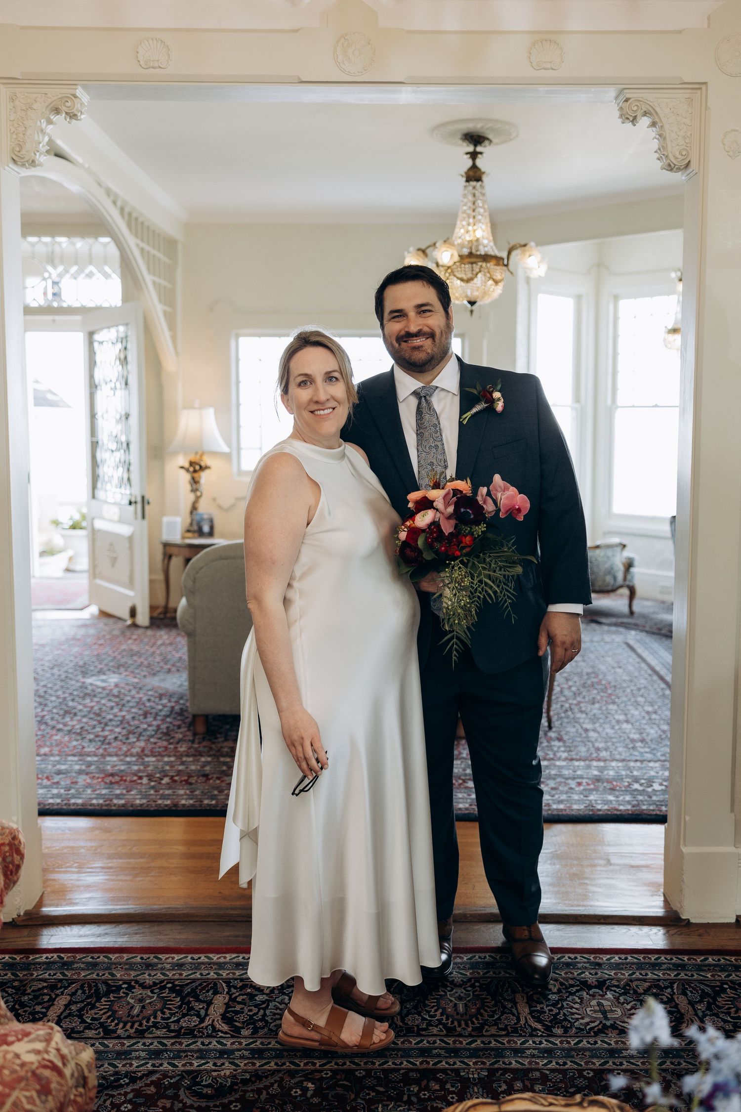 A couple stands together in a bright, elegant room with ornate carpeting and chandeliers during their intimate wedding ceremony.