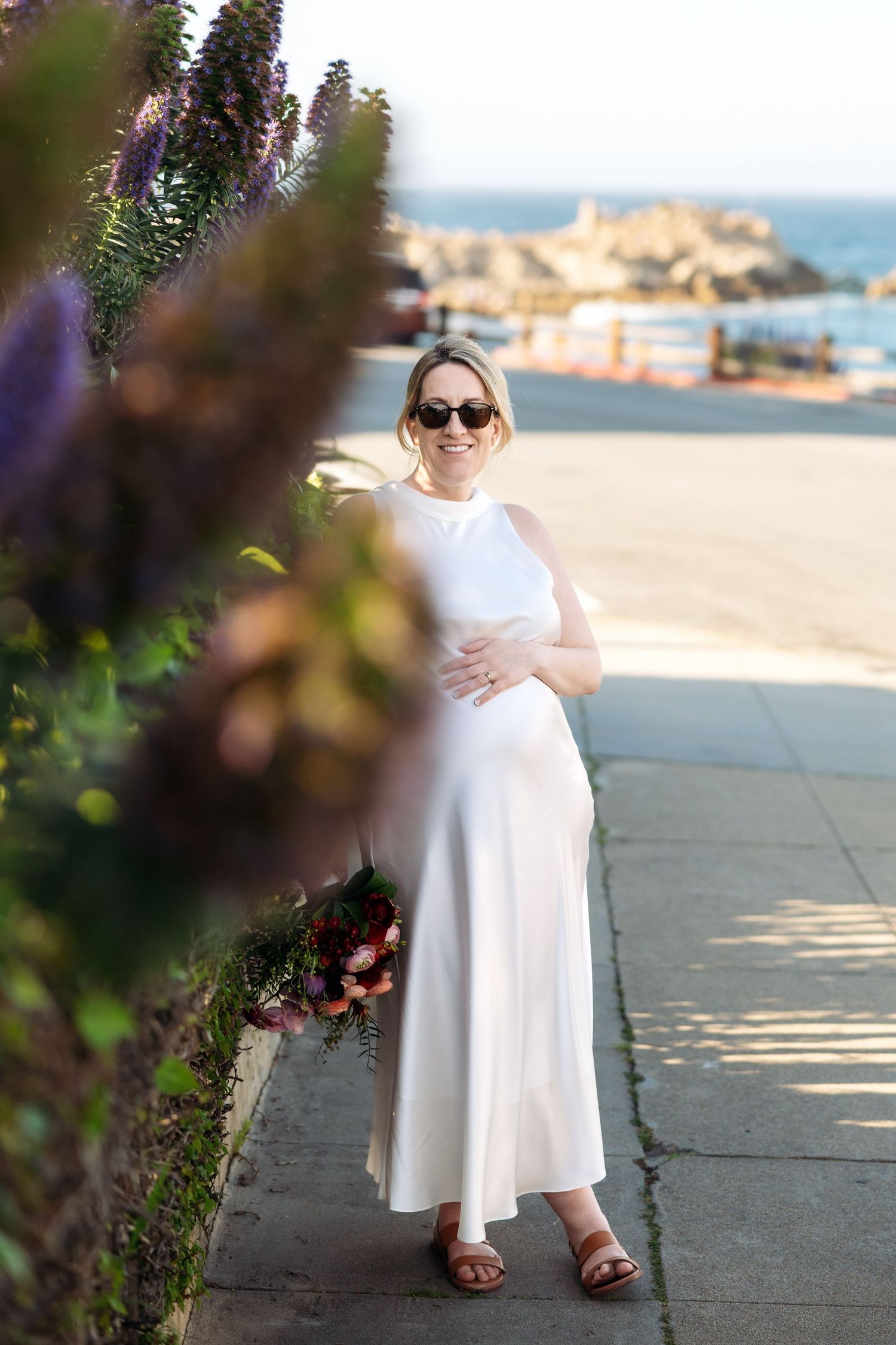 A bride in a white dress and sunglasses poses against flowering bushes near a coastal backdrop.