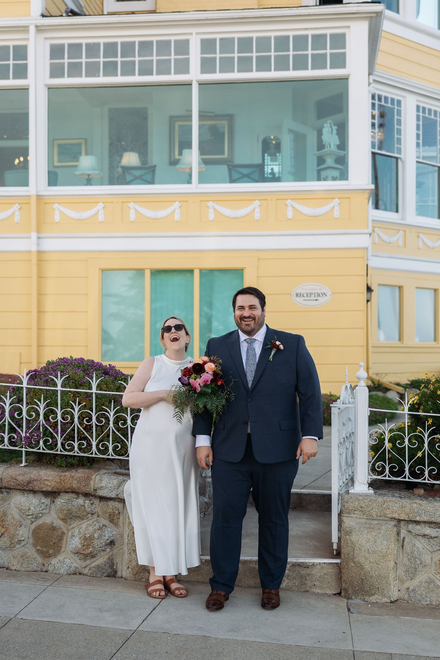 A couple in wedding attire stands in front of a yellow Victorian-style building with ornate windows and stone fence.