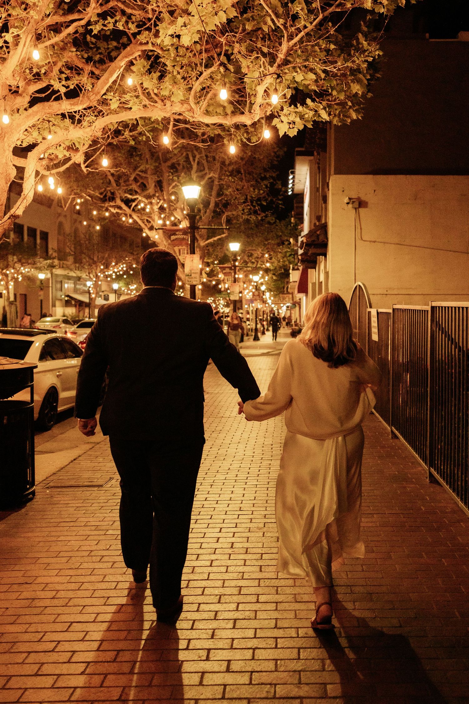 A couple walks hand in hand down a romantic street lit by warm glowing string lights in trees.