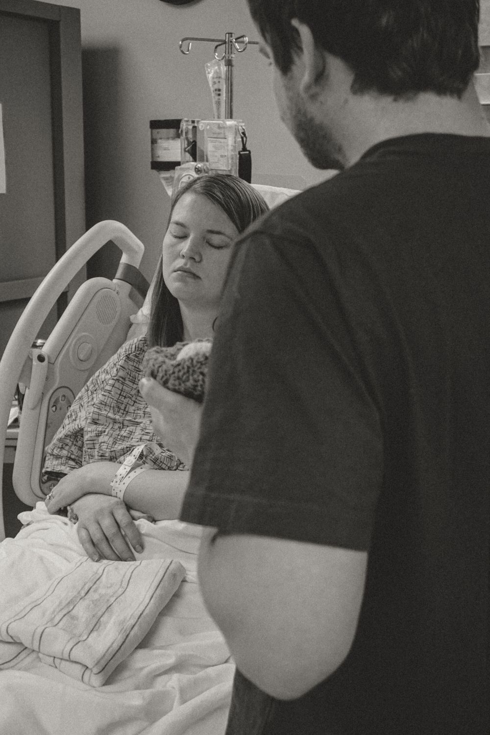 Black and white photo showing a patient resting in a hospital bed with medical equipment and a visitor standing nearby.